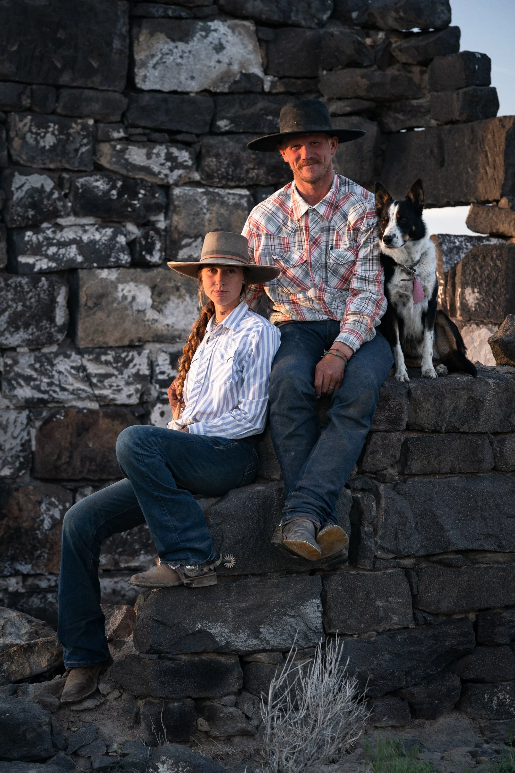 Buckaroo couple seated on a stone wall with a black-and-white herding dog, wearing cowboy hats, plaid shirts, and jeans.