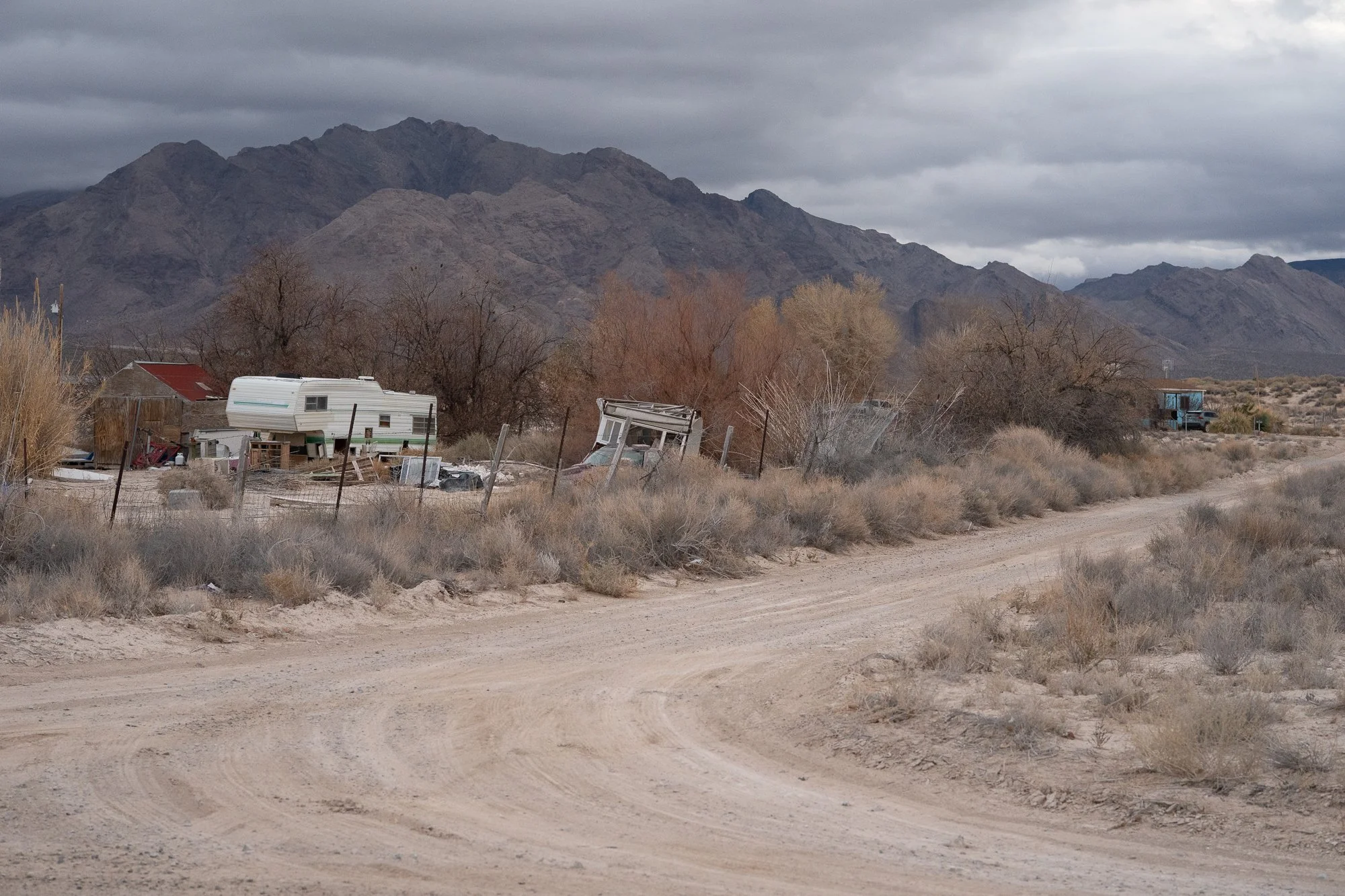 Old travel trailers and scattered debris along a dirt road in a rural Nevada desert community, with bare trees and a large mountain range under gray clouds.