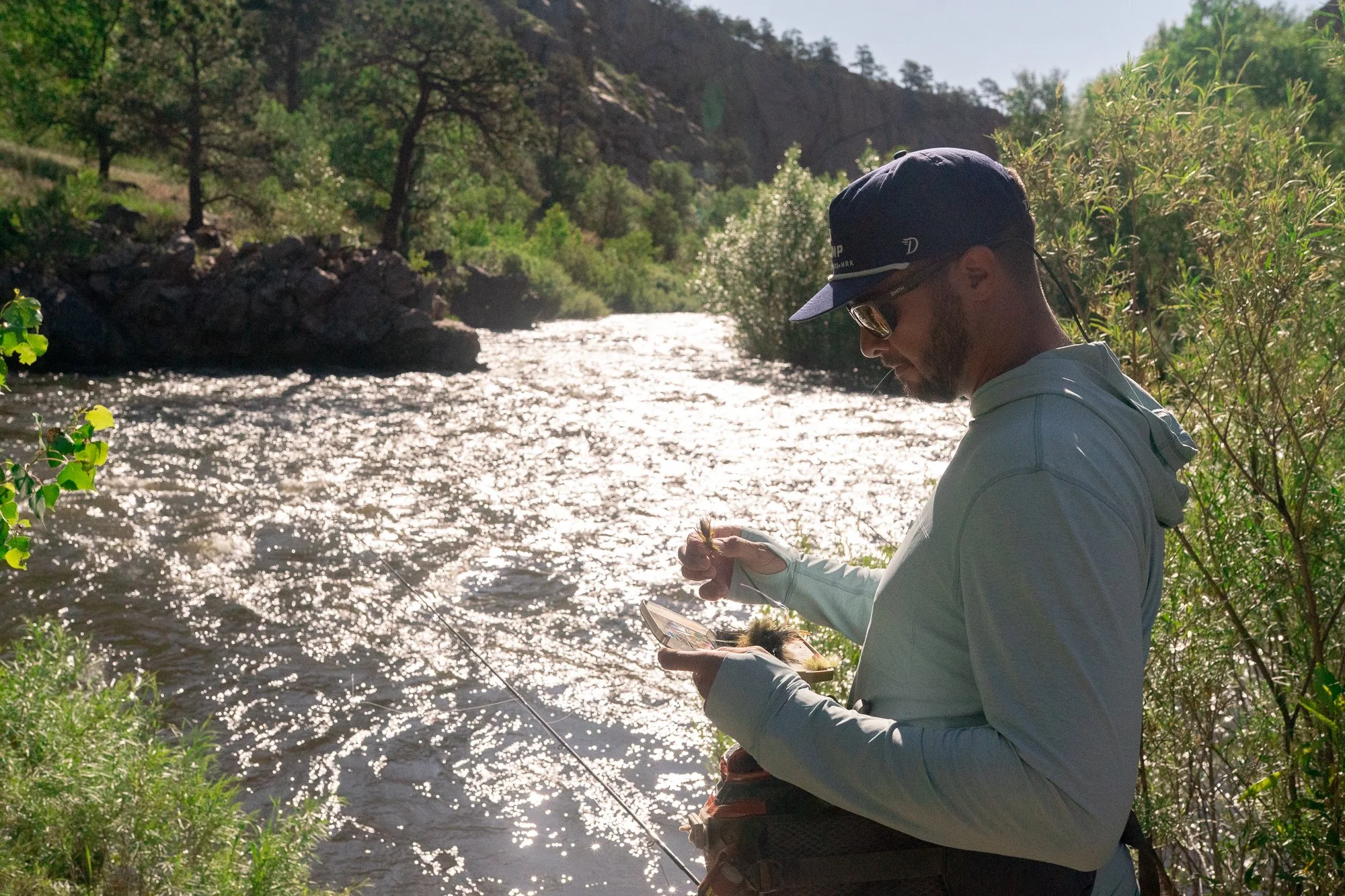 An angler in Duck Camp apparel selects a fly beside the river with a fly rod nearby