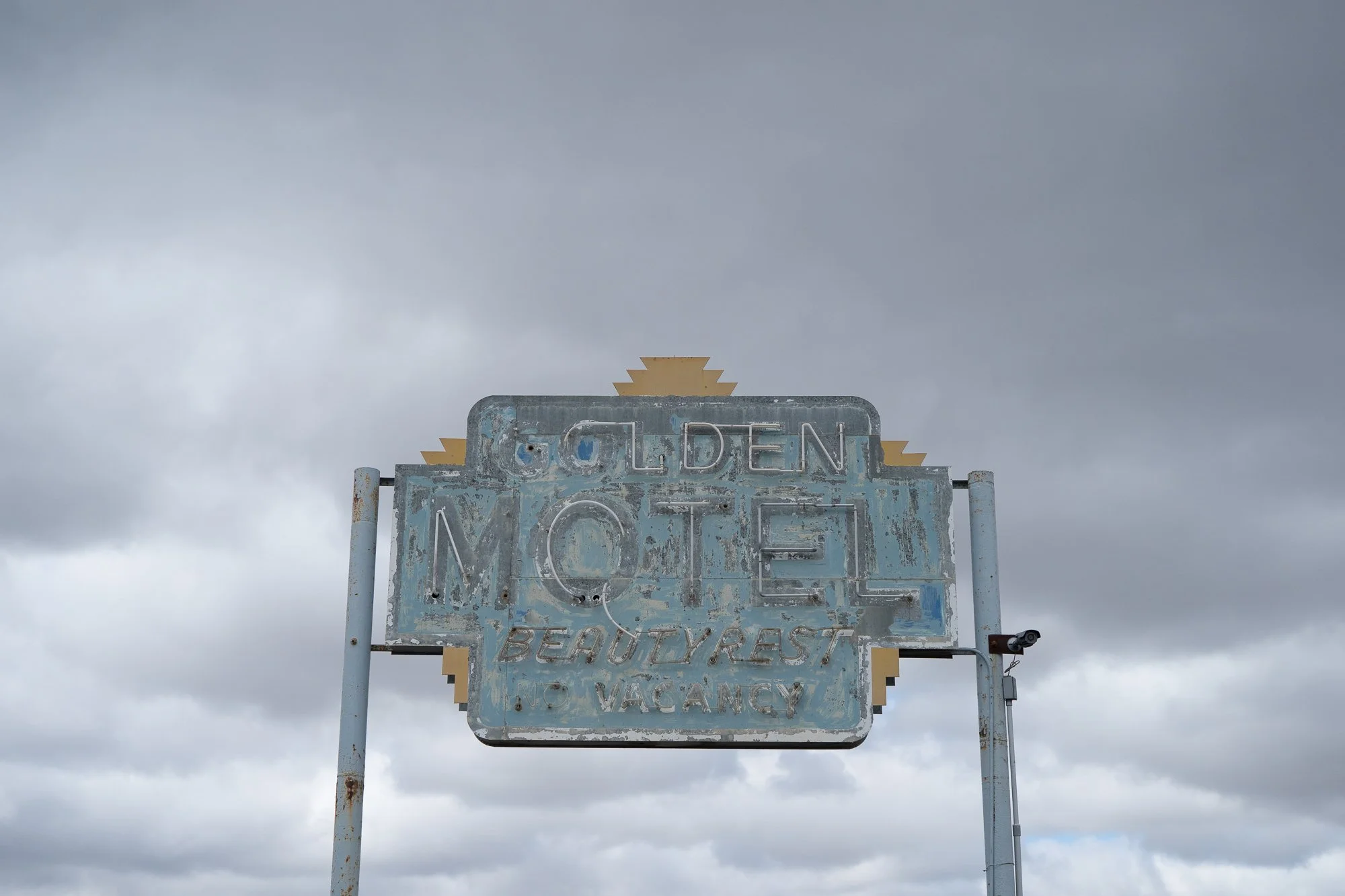 Weathered light blue Golden Motel neon sign with heavily peeling paint, mounted on metal poles against a gray overcast sky in rural Nevada.