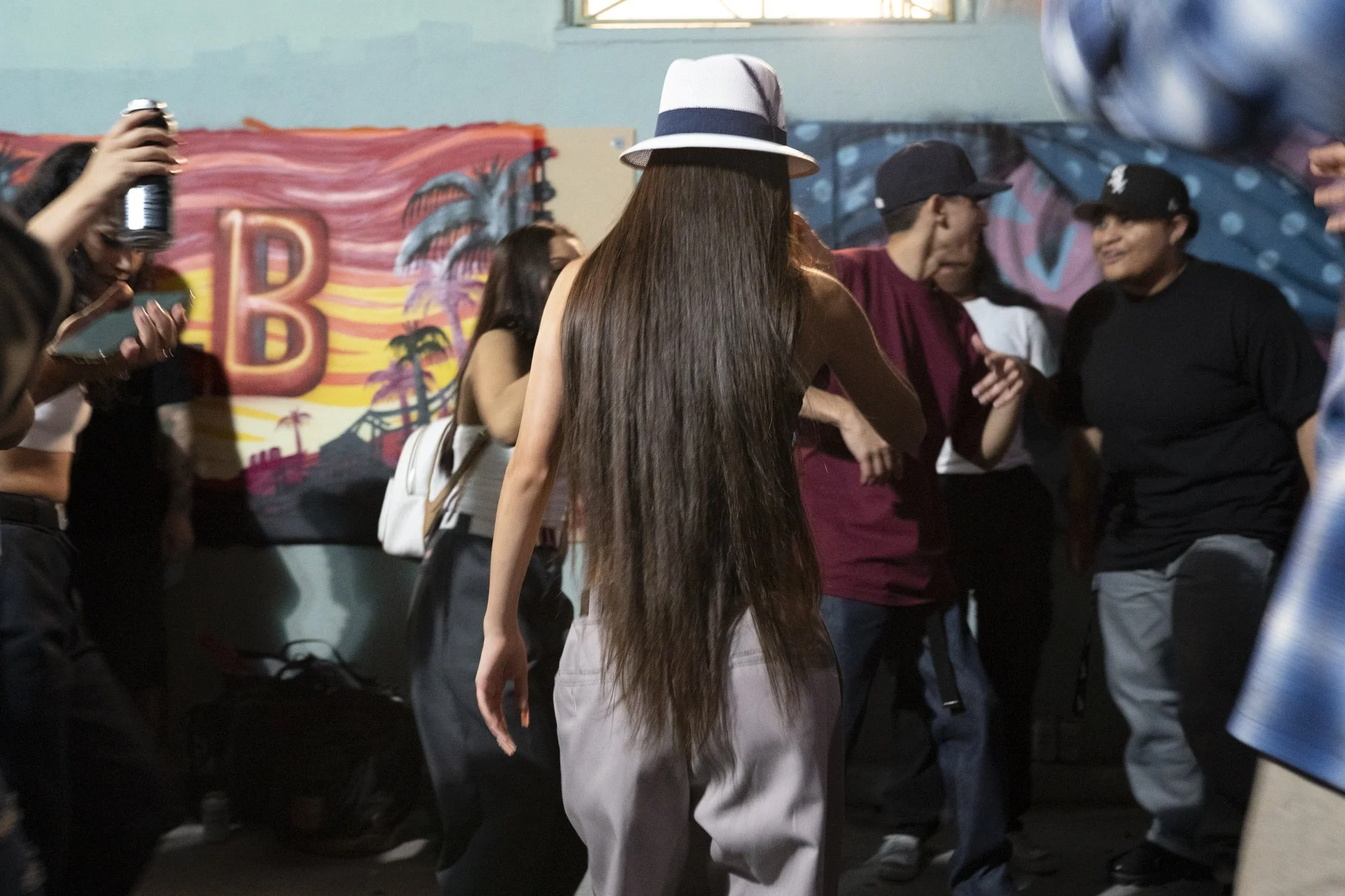 Woman in a wide-brimmed hat dancing in front of a Long Beach mural at the Syndicate Barbershop alley party with a crowd of people dancing and filming around her