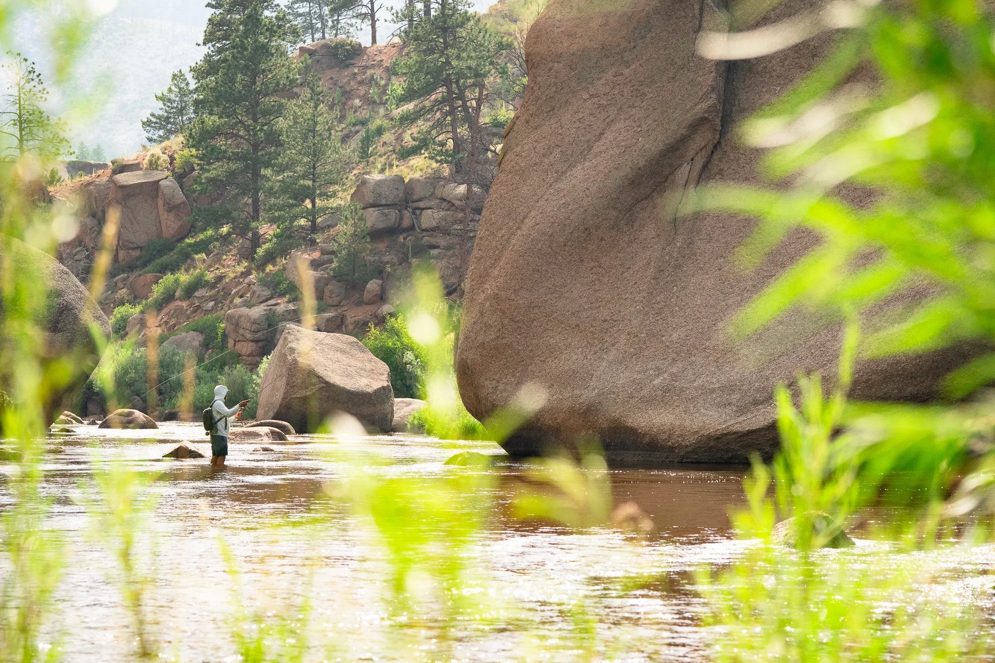 A fly angler stands in the river near large boulders with a rod in hand
