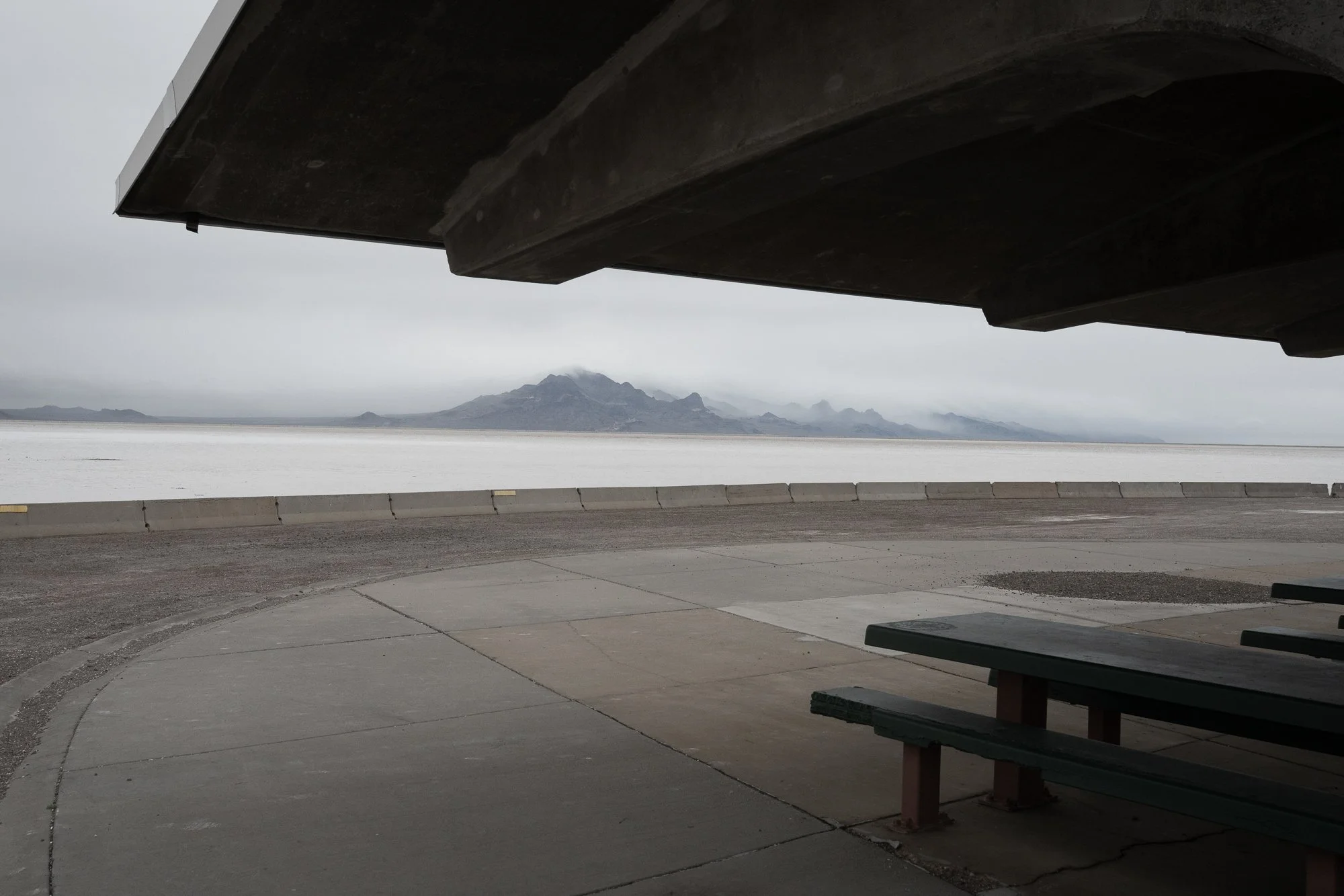 A roadside overlook with concrete pavement and picnic tables faces a broad salt flat and distant mountain range beneath an overcast sky.