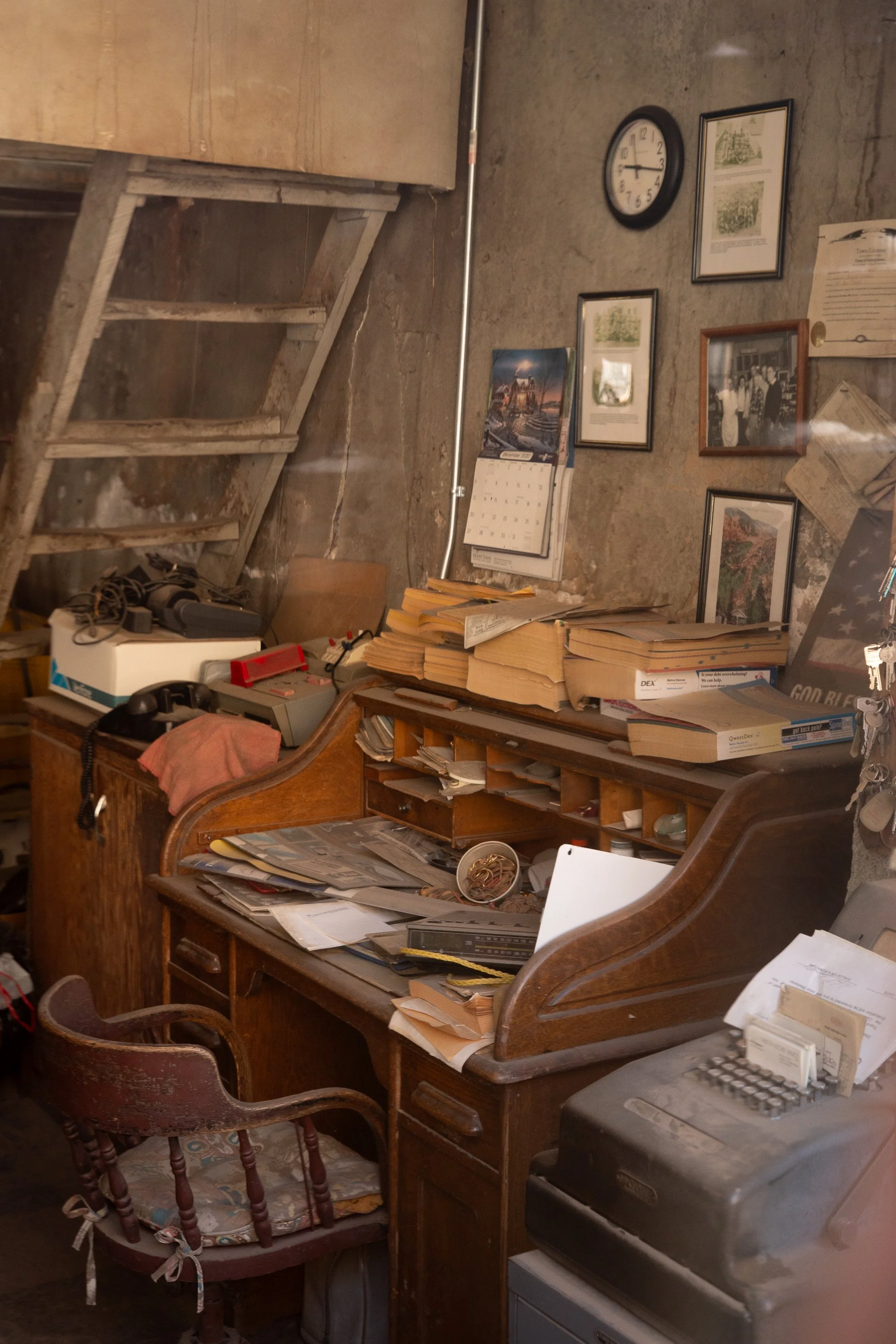  A color photograph of an office desk positioned beneath a staircase, surrounded by papers, books, and framed photographs. This image documents an American interior workspace and the accumulation of everyday objects over time - Georgetown, Colorado -