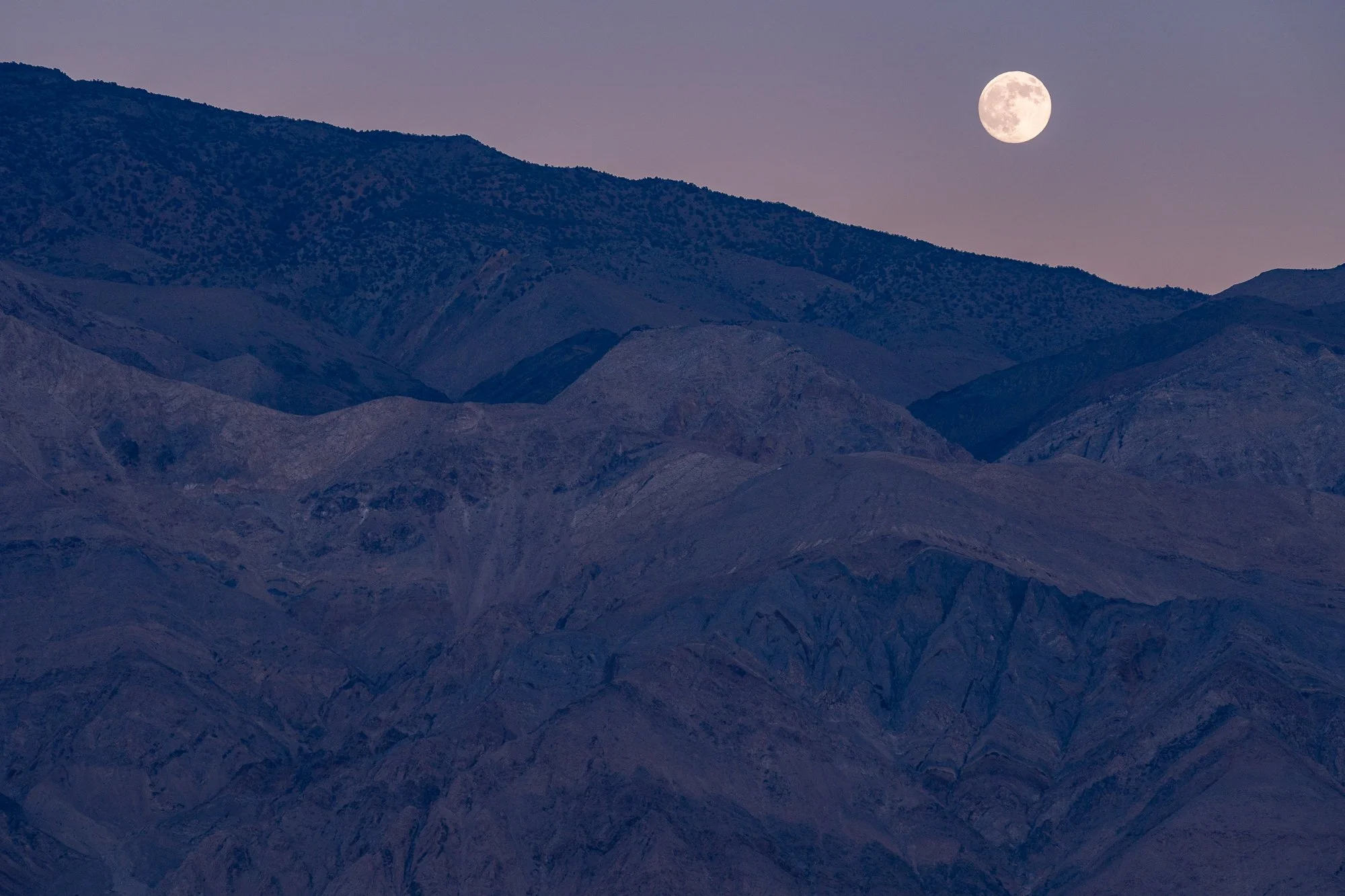  A full moon hangs above layered desert mountains at dusk, the landscape settling into deep blue tones as daylight fades. The photograph balances scale, stillness, and the quiet presence of time passing. 