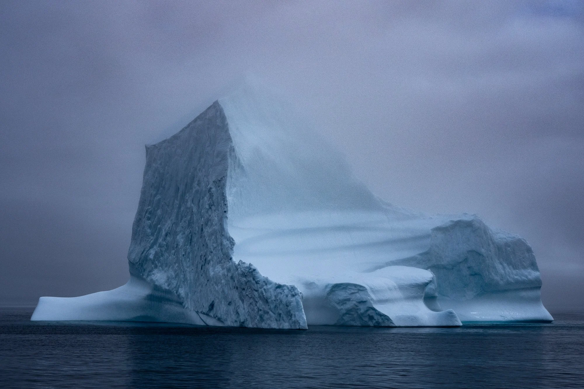  A monumental iceberg drifts through calm, dark water beneath heavy cloud cover. Subtle tonal shifts reveal texture and form, emphasizing isolation, impermanence, and the slow movement of the natural world. 