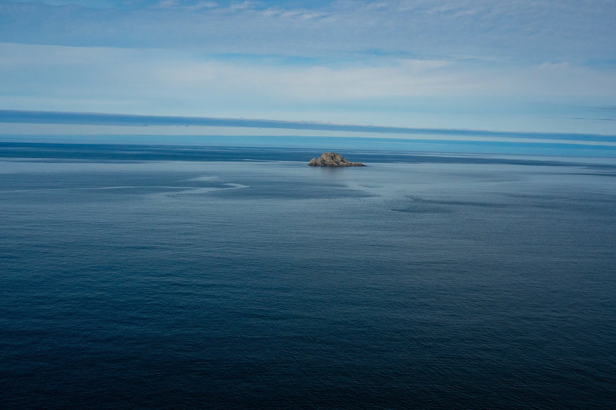  A small rocky island sits alone within a wide expanse of calm water and layered sky. The minimal composition invites contemplation, scale, and a sense of distance without destination. 