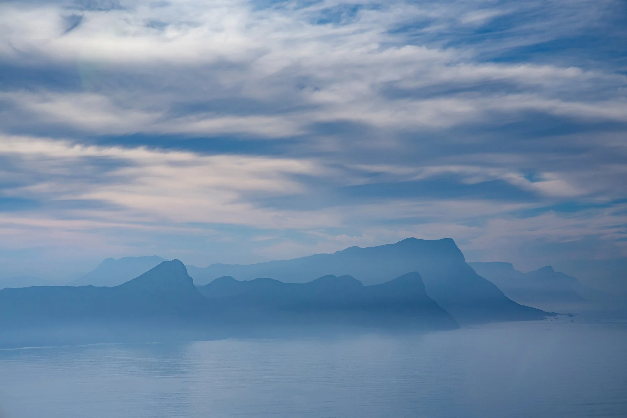  Soft layers of coastal mountains recede into the distance beneath a calm, cloud-filled sky. The muted blue tones and gentle atmospheric haze create a sense of depth, quiet, and visual balance, emphasizing stillness and the subtle rhythms of land, wa