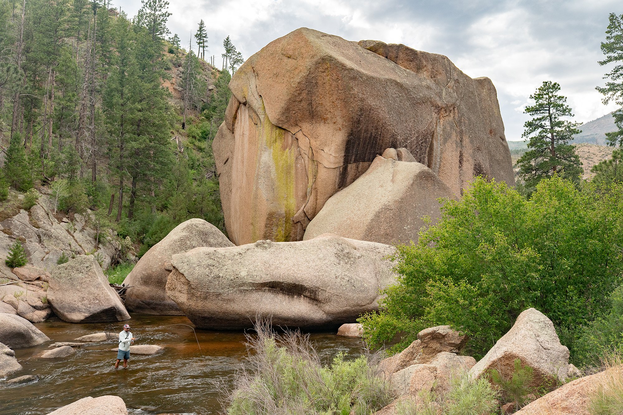  Photograph of a fly fisherman casting into a beautiful section of the South Platter River surrounded by enourmous boulders. 