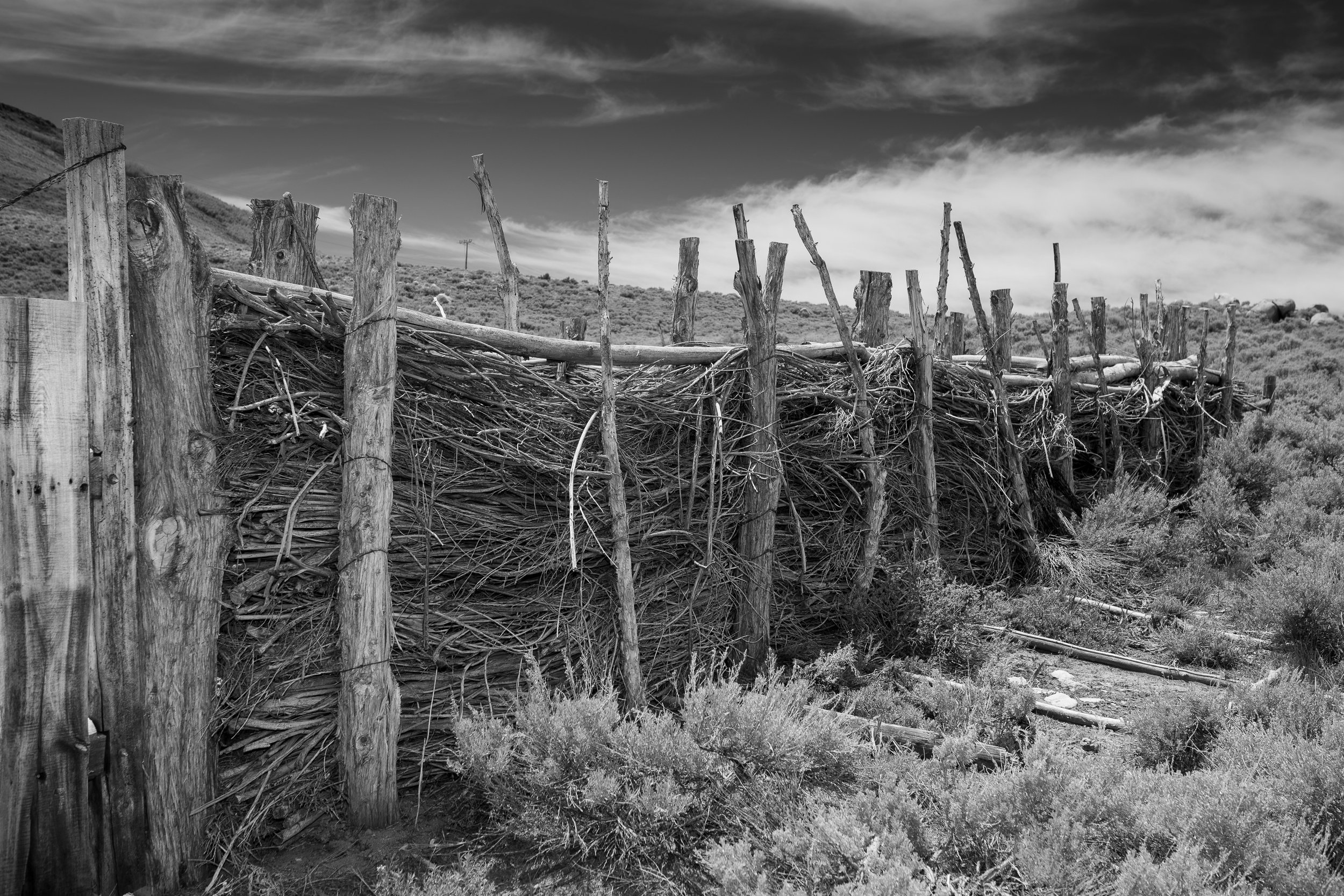  Hand-built corral wall woven from branches and posts in a high desert landscape under dramatic skies. 