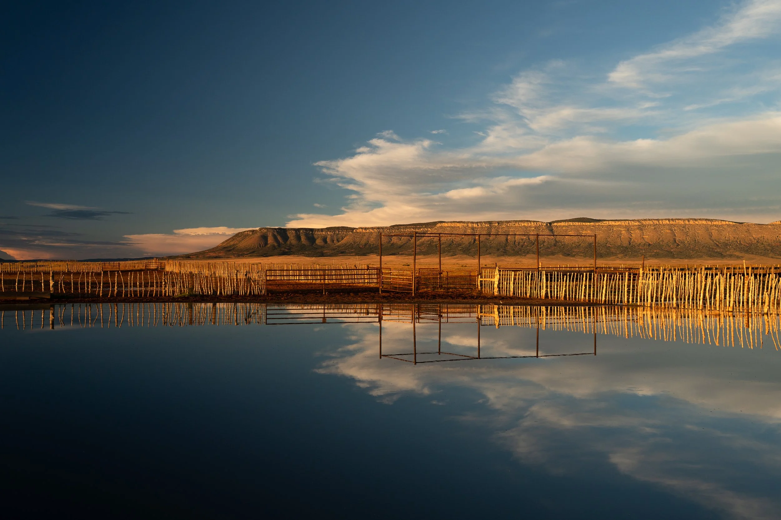  Golden light hits a rustic corral beside still water reflecting clouds and long wooden fencing in New Mexico. 