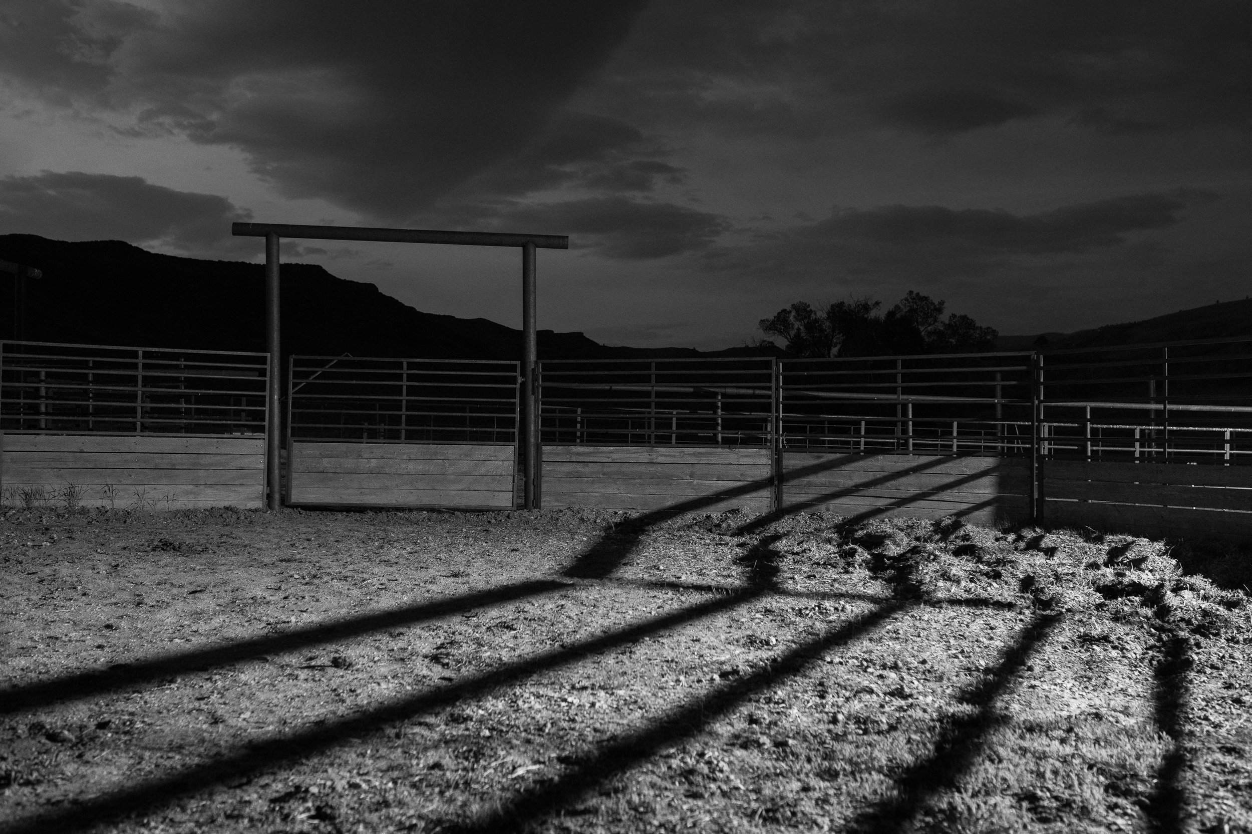  High contrast black and white image of corral fencing casting long shadows across the ground at night. 