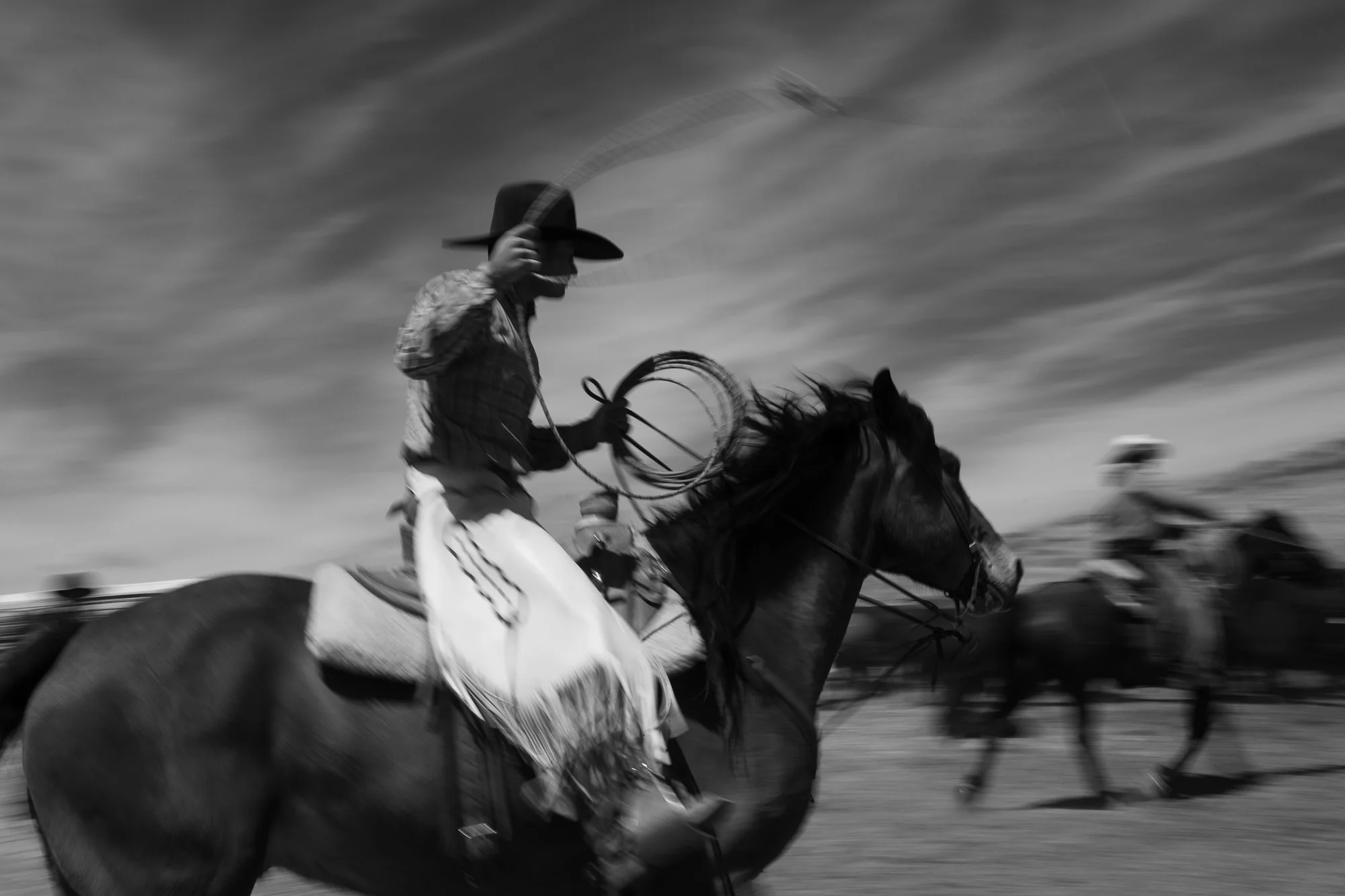  Black and white photograph of a cowboy swinging a rope from horseback, photographed with motion blur. 