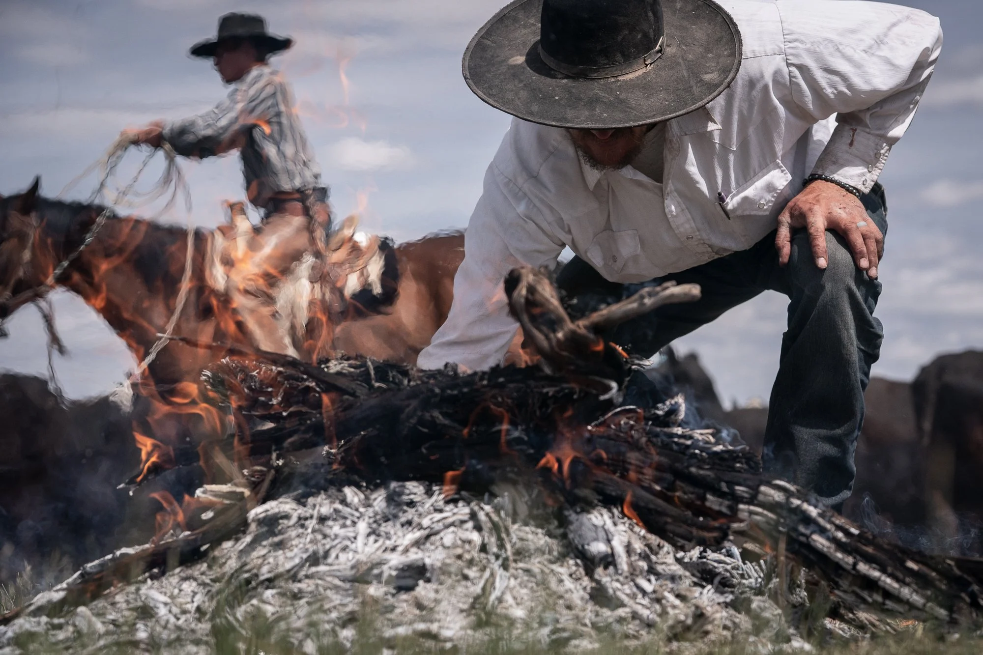 Cowboy tending a branding fire as another rider works cattle from horseback during a traditional ranch branding. 
