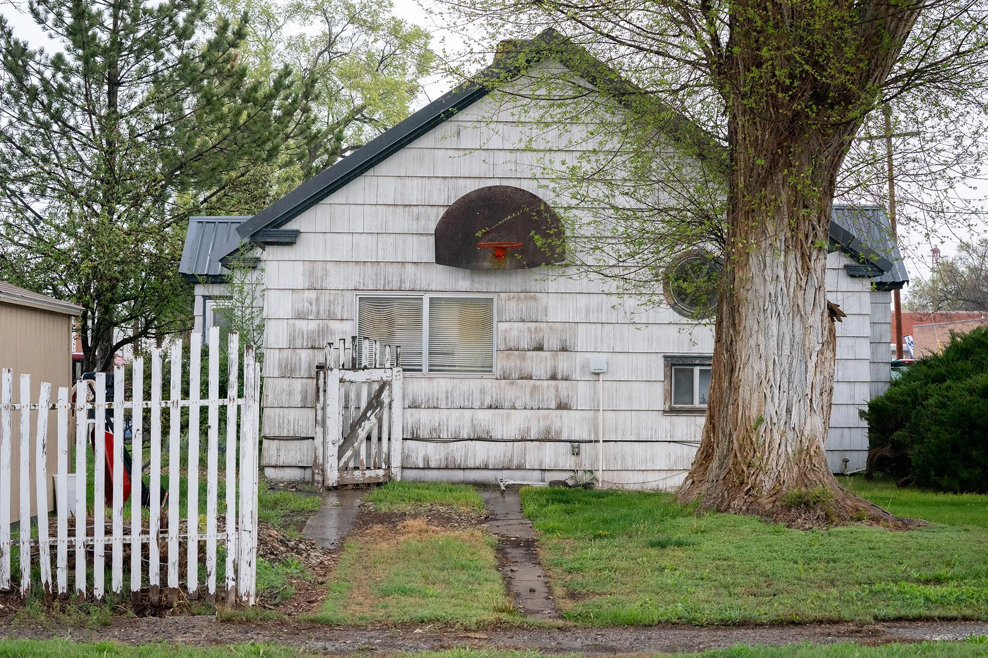  Photograph of a basketball hoop hanging on the side of an old house in a small American town.  