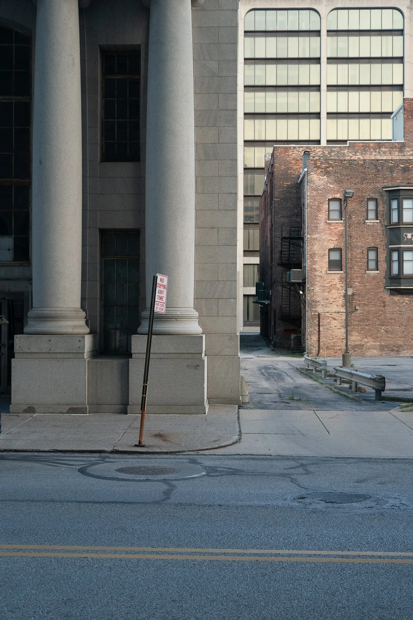 City street lined with brick buildings and large stone columns, with utility poles and a no stopping sign at the curb.
