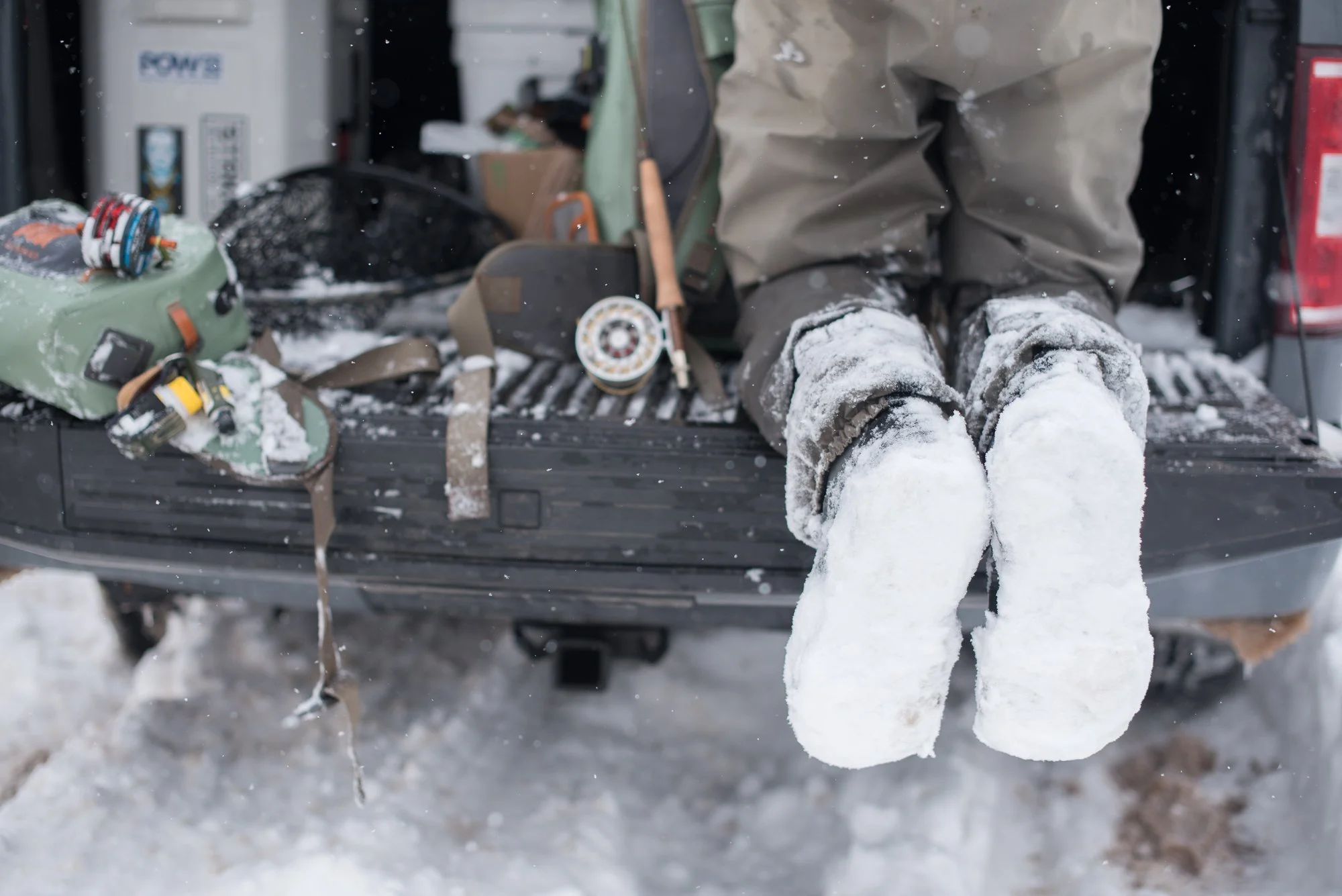 A fisherman sits on a truck tailgate with snow-covered boots and fishing gear after time on Clear Creek.