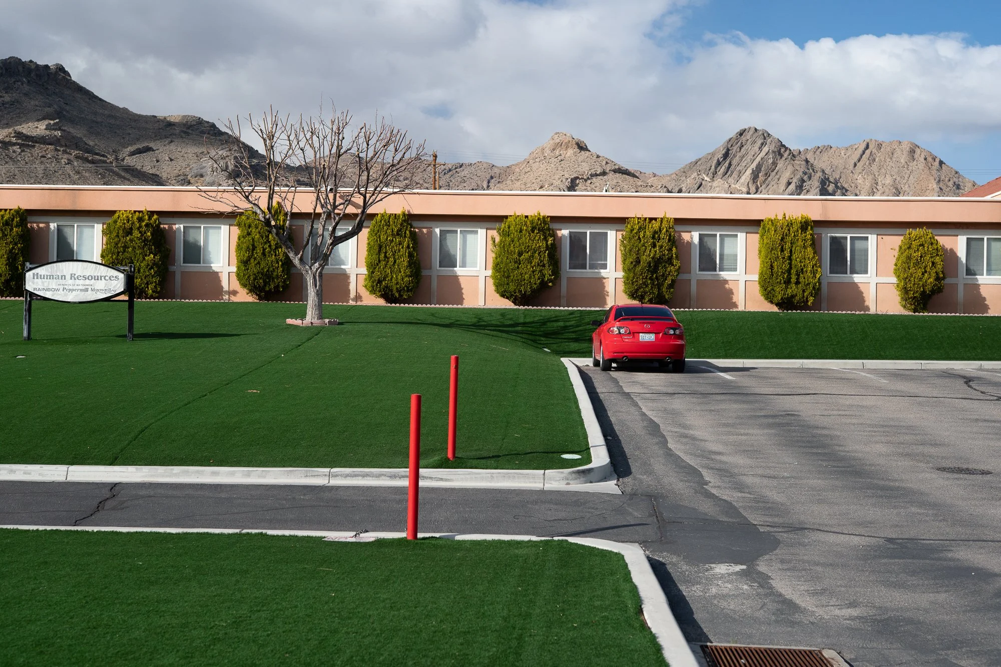 Single-story Nevada office building with artificial turf, trimmed shrubs, and a red car parked near the entrance, with jagged desert mountains rising directly behind it.