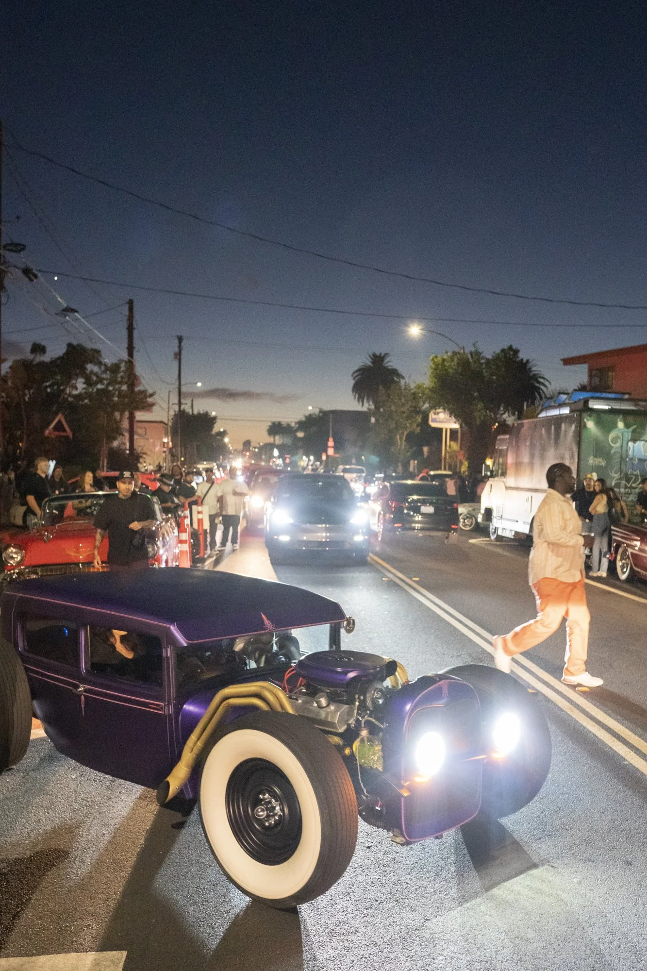 Matte purple chopped hot rod parked on a Long Beach street at dusk with a crowd lining the sidewalk and palm trees visible against the evening sky