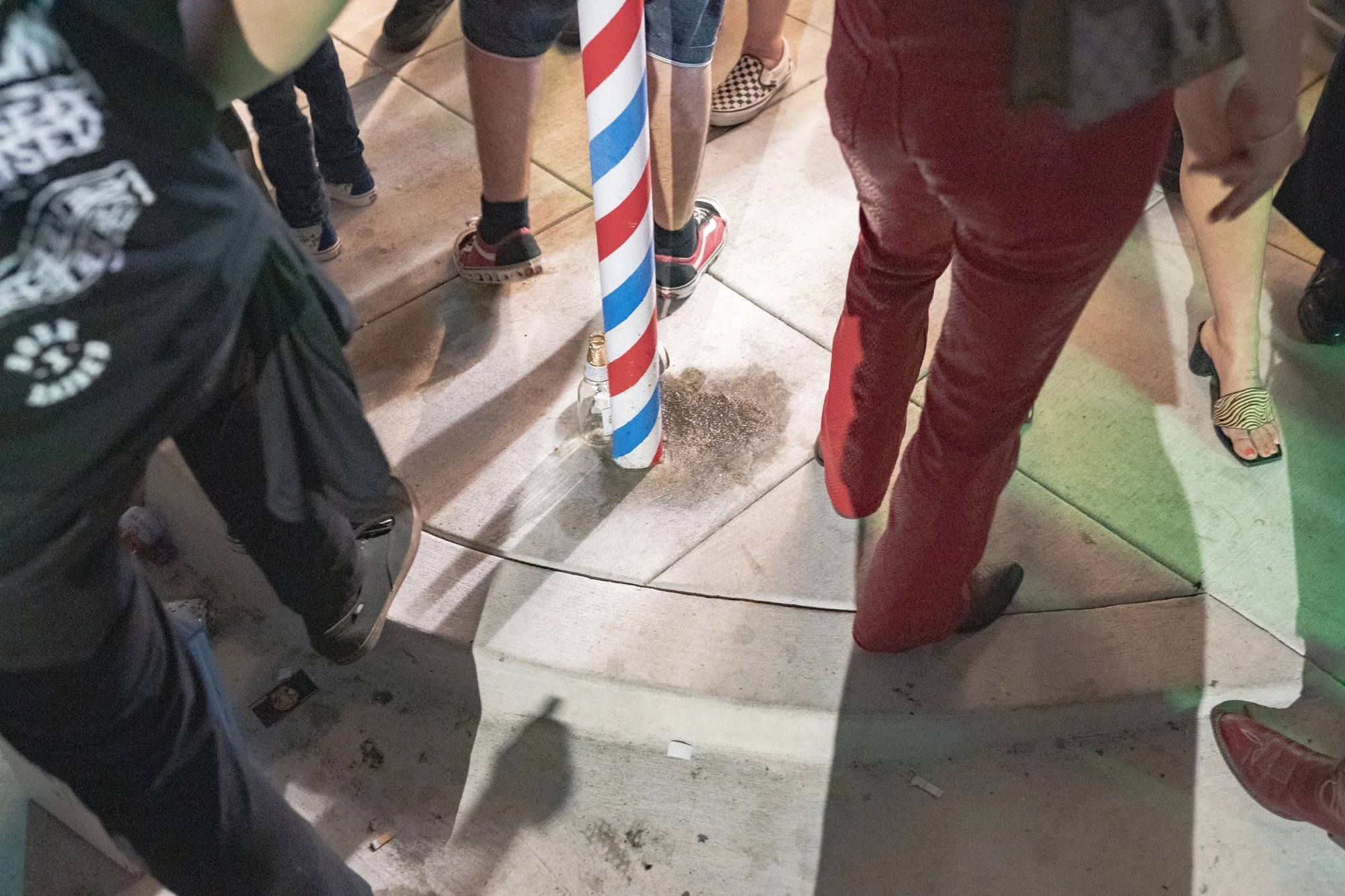 Ground-level view of a barber pole base surrounded by crowd feet in mixed footwear on the sidewalk outside Syndicate Barbershop at night