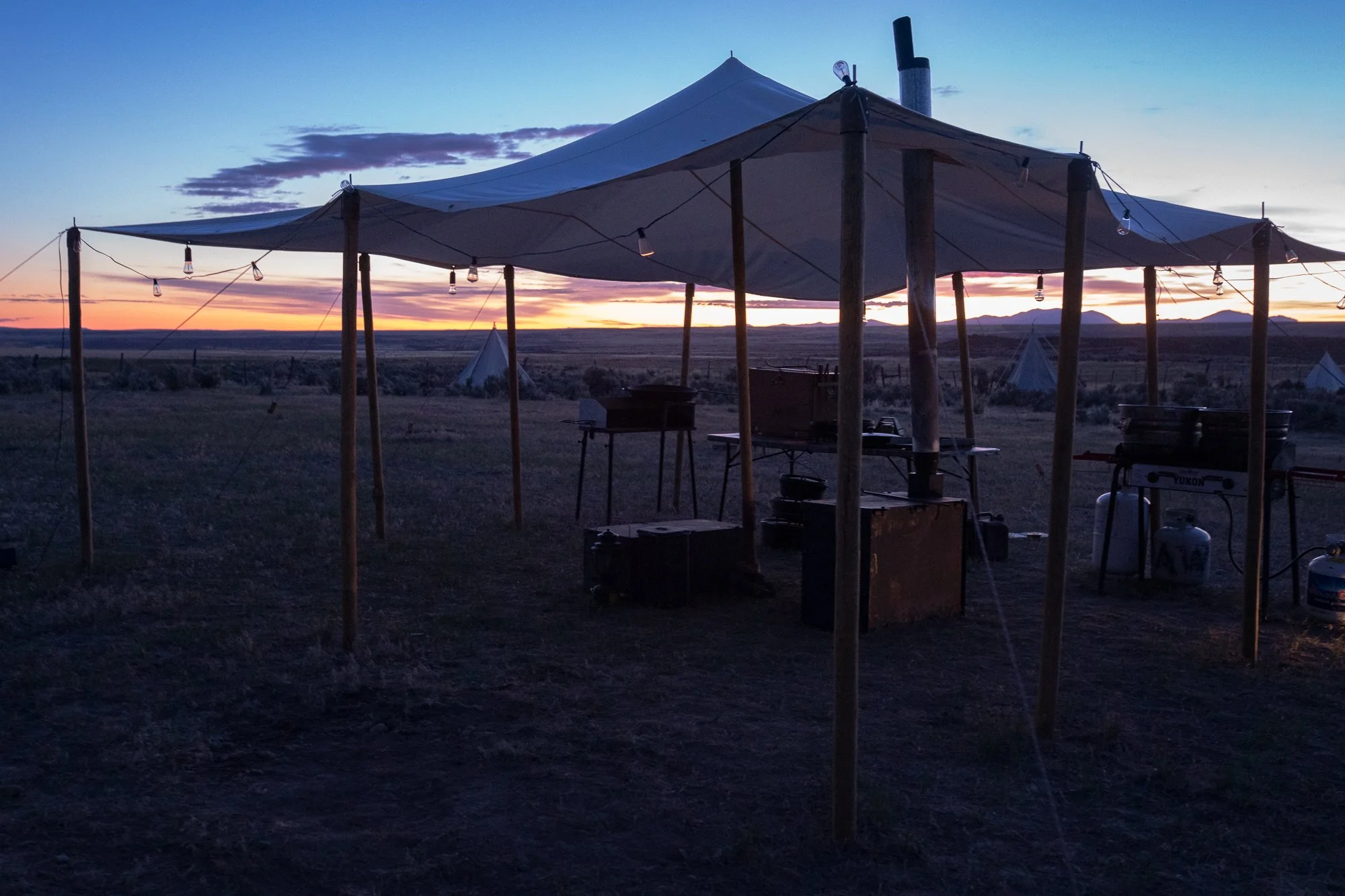 Primitive stoves are set up under a chuckwagon tent with cowboy teepees in the background at sunrise on a working cattle ranch