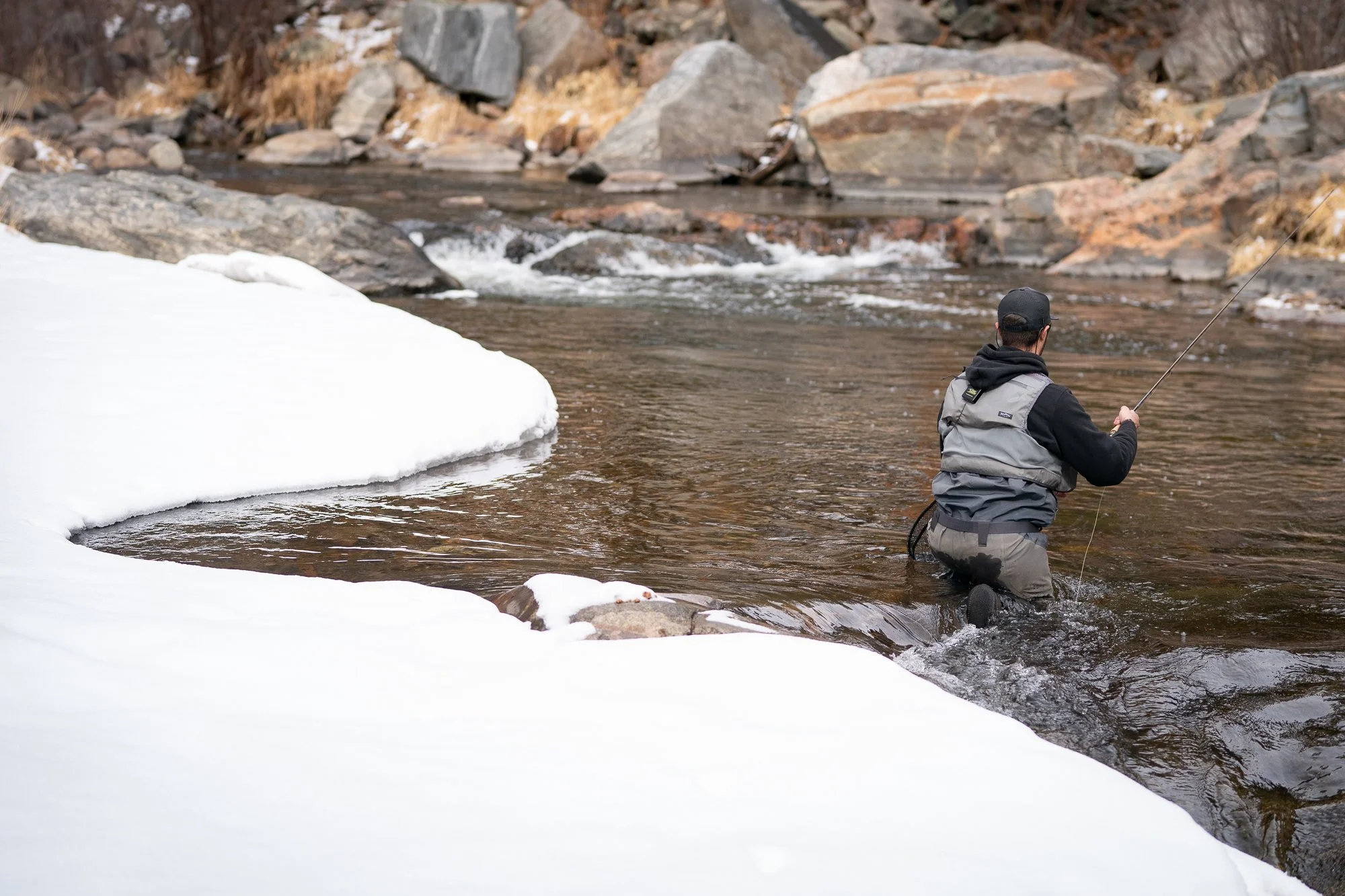 A fly fisherman kneels in a stream next to snow covered banks to avoid spooking fish