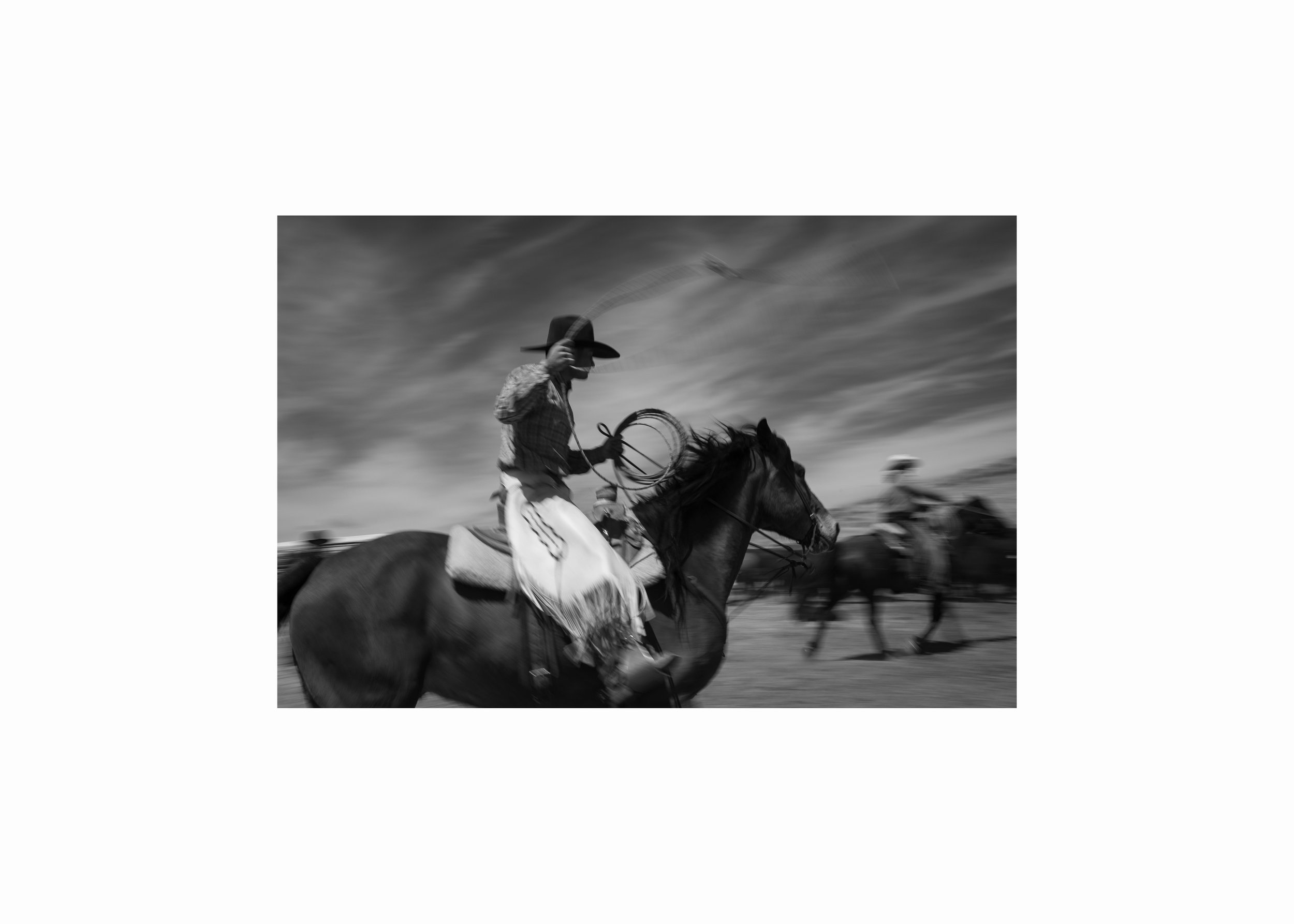 Black and white photograph of a cowboy on horseback swinging a rope, captured with motion blur while working cattle.