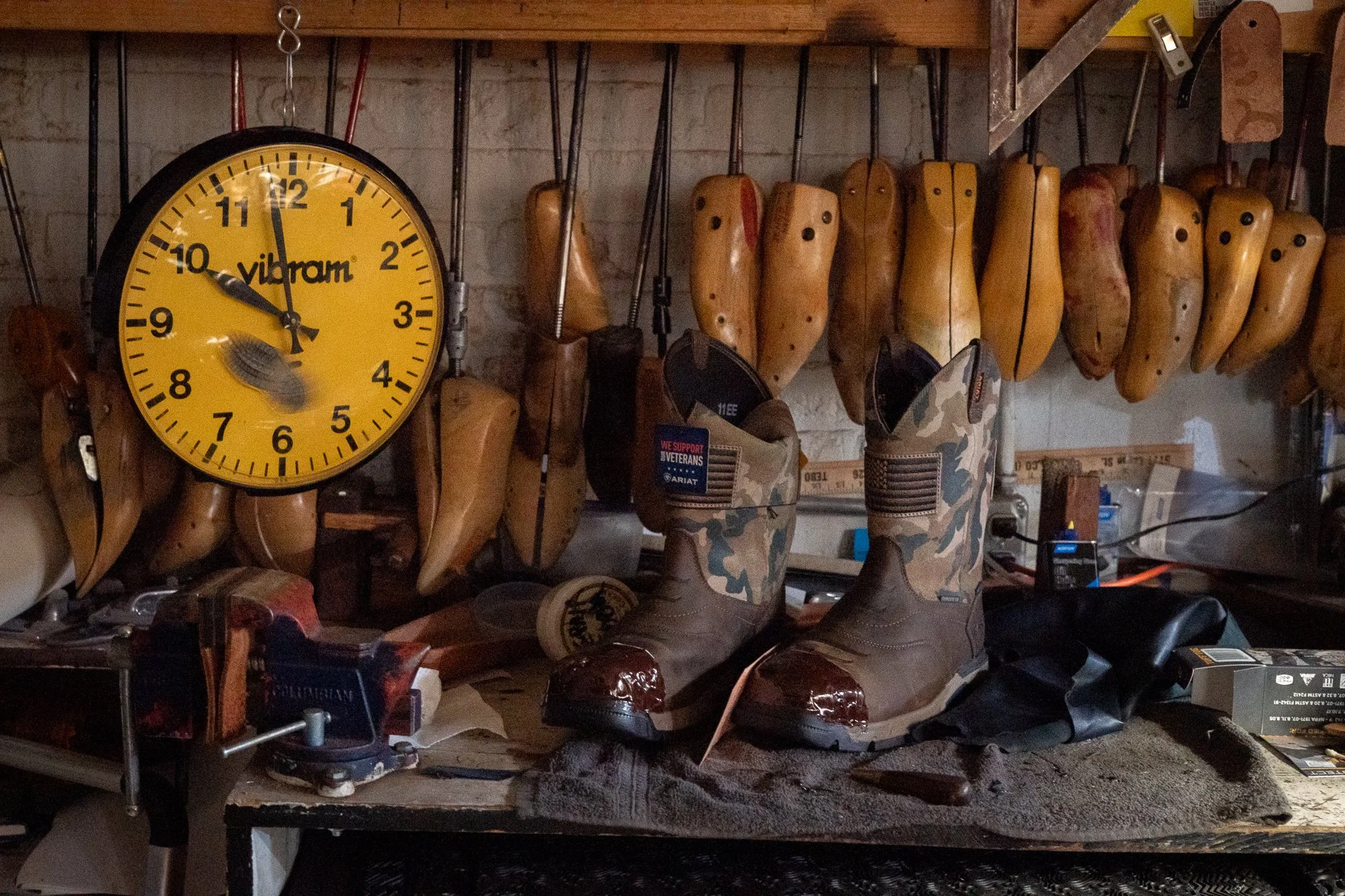 Camouflage-patterned cowboy boots on a repair bench beneath wooden boot lasts and a yellow wall clock at City Shoe & Saddle Shop in Douglas, Wyoming.