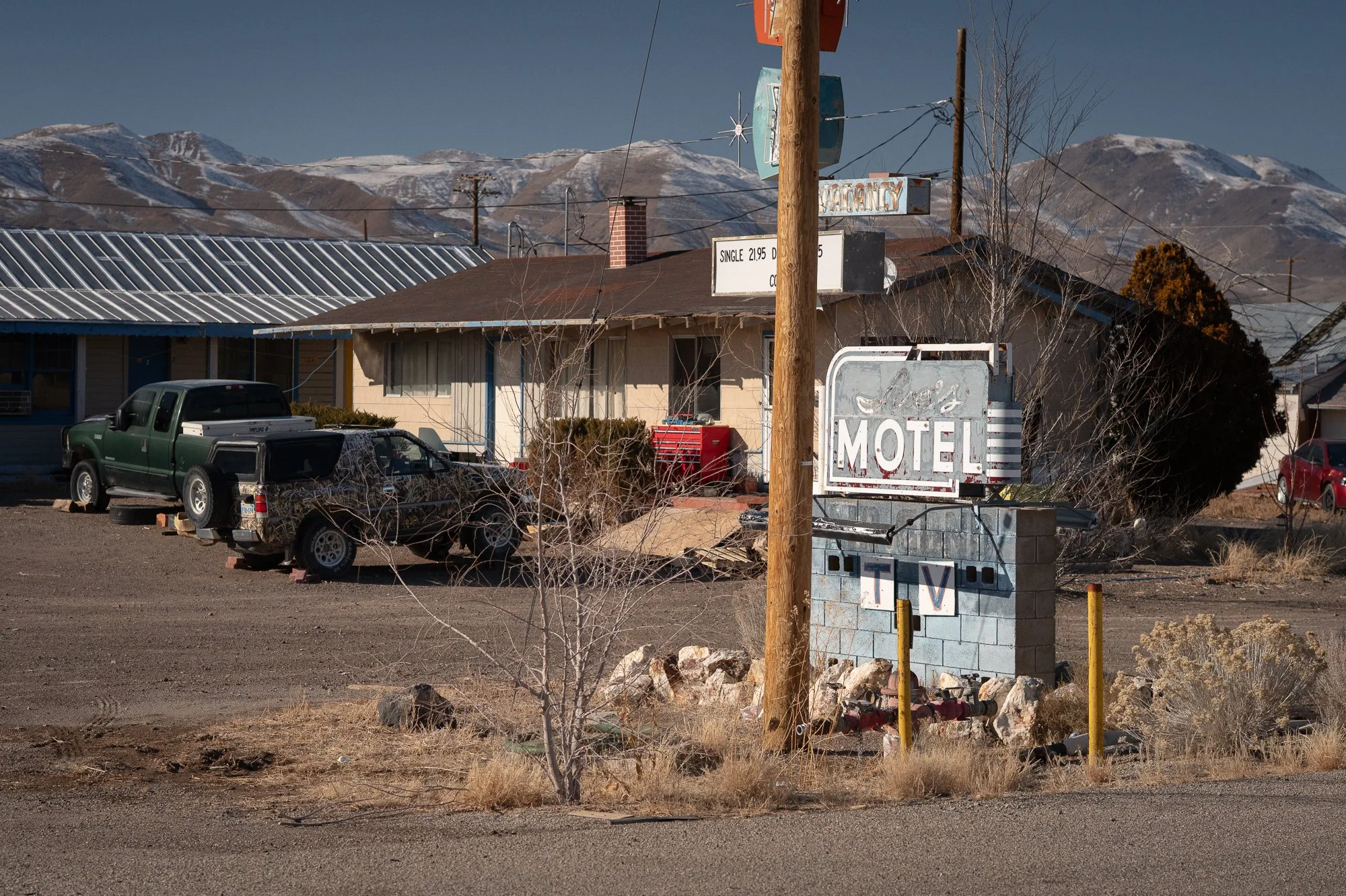 A roadside motel with a large motel sign, parked trucks, utility poles, dry grass, and snow-covered mountains in the background.