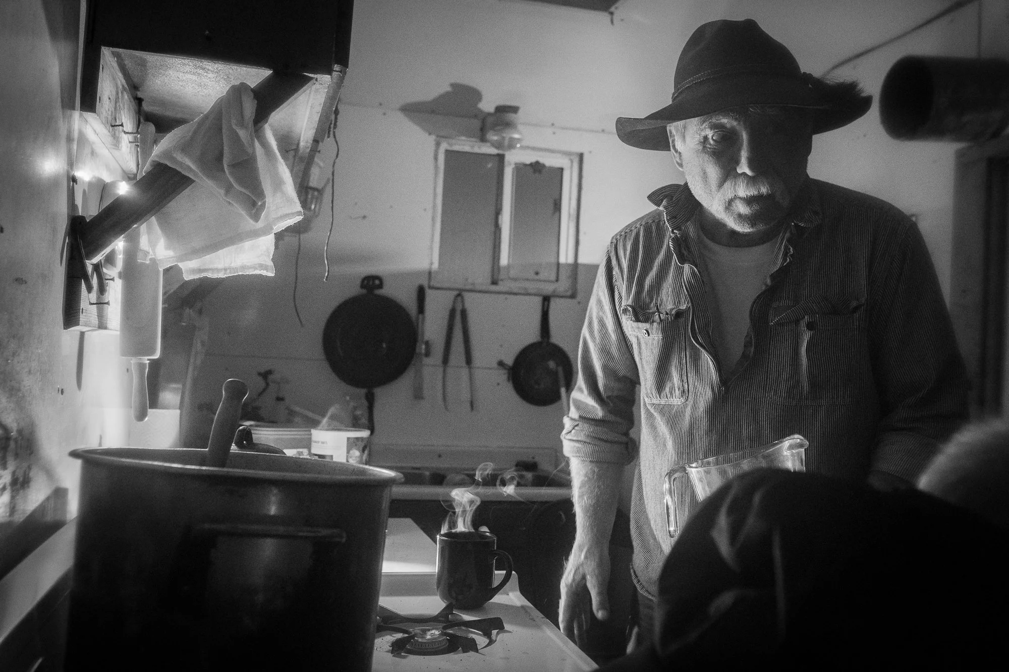 Black-and-white photograph of a cowboy in a hat and work shirt standing beside a steaming mug and cooking pot in a ranch kitchen.