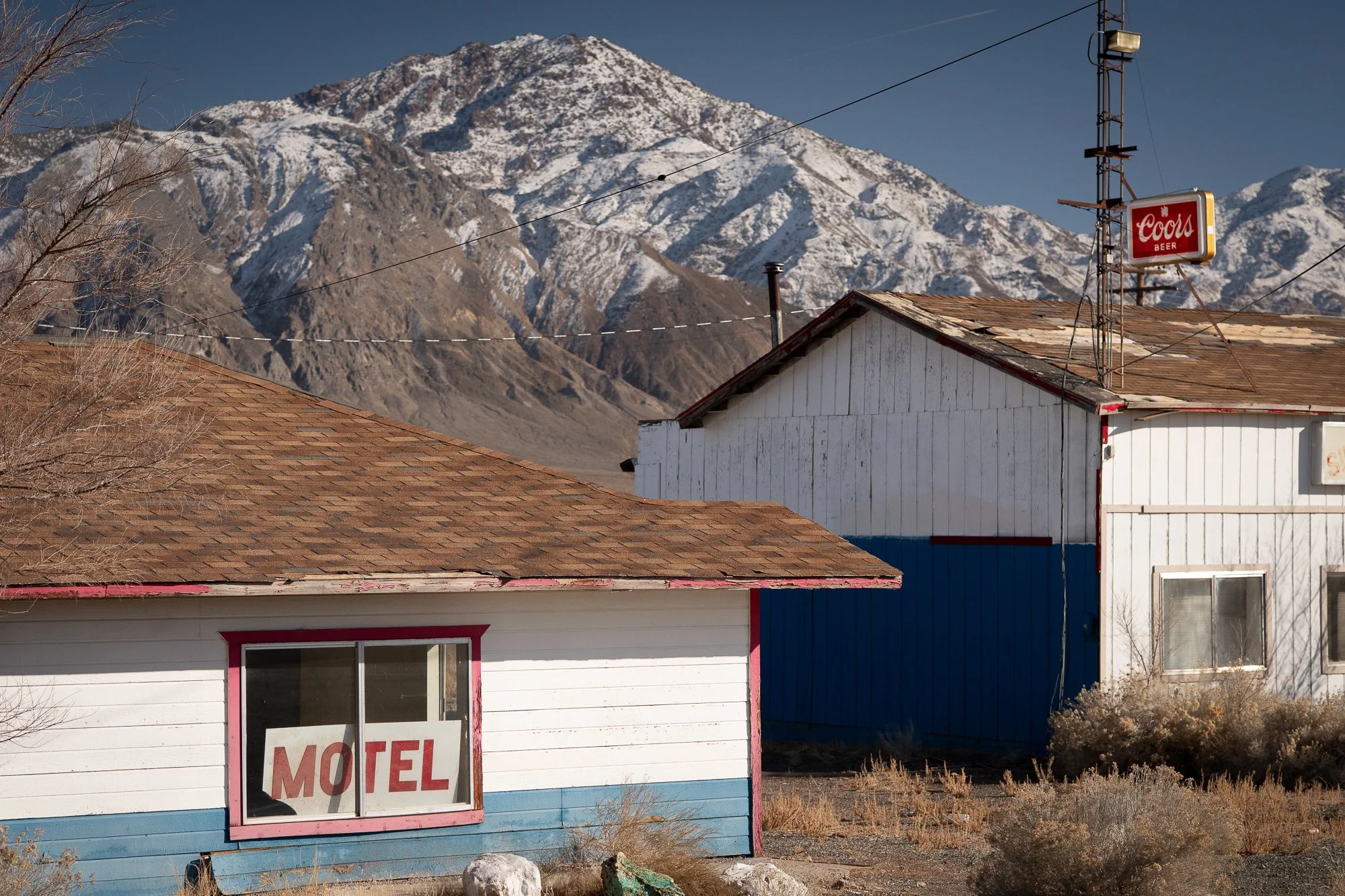 Small Nevada roadside motel with a hand-lettered Motel sign in the window, a Coors Beer sign on a post nearby, and a snow-covered mountain visible directly behind the building.