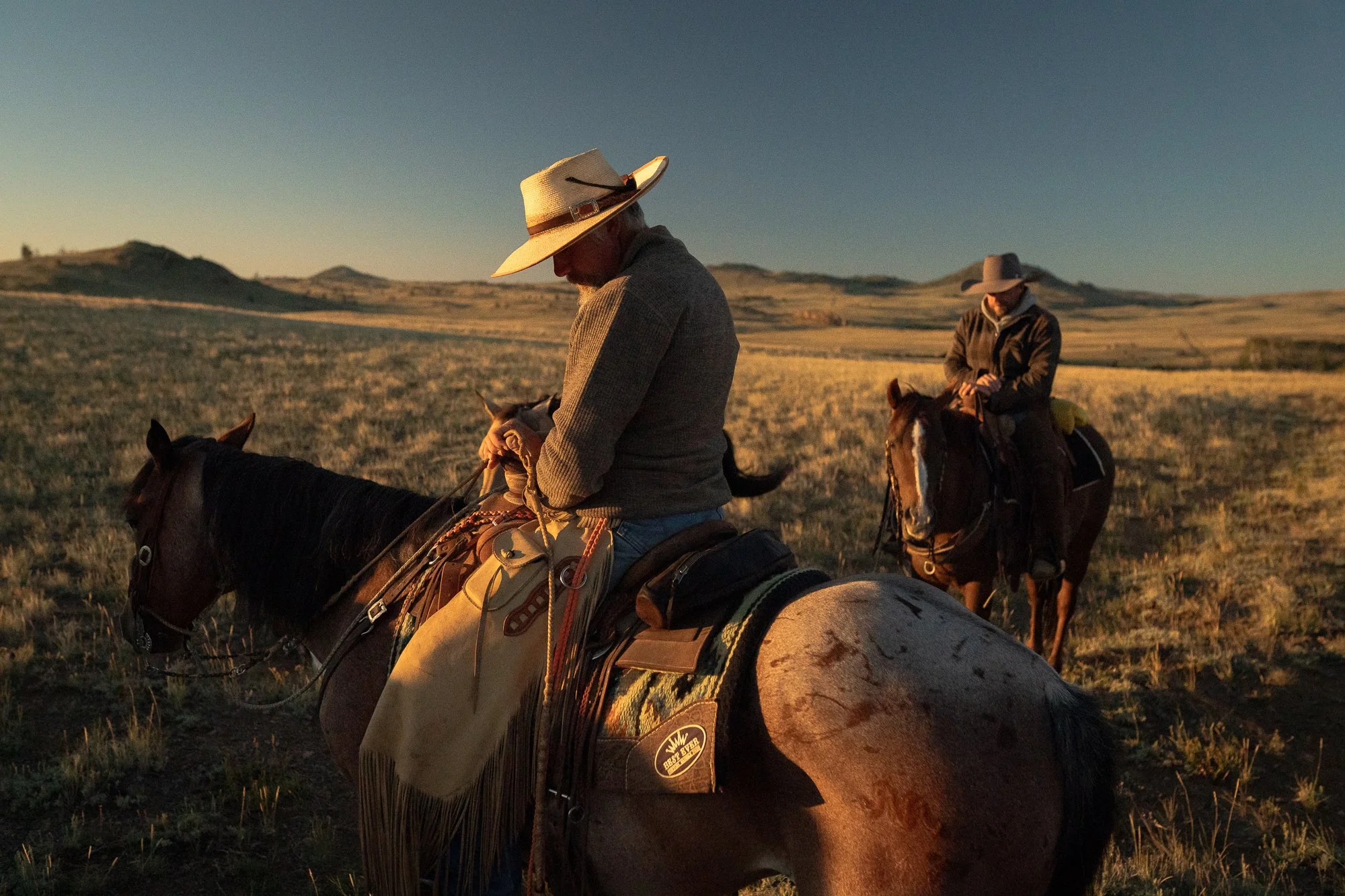 Cowboys riding horses across open range at golden hour with long shadows