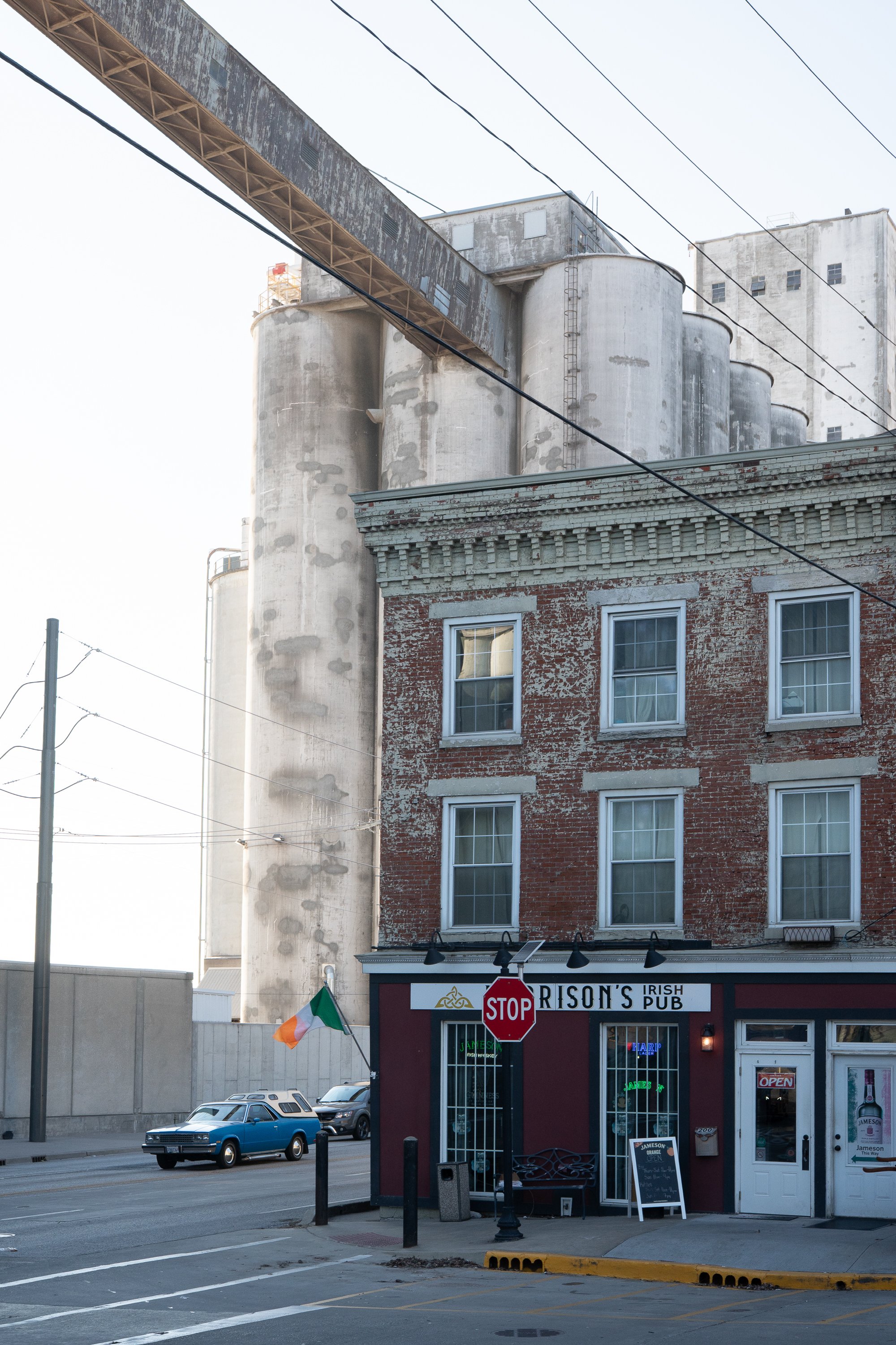 Brick Irish pub storefront beneath large concrete grain silos, with overhead utility lines, parked cars, and a stop sign along the street.