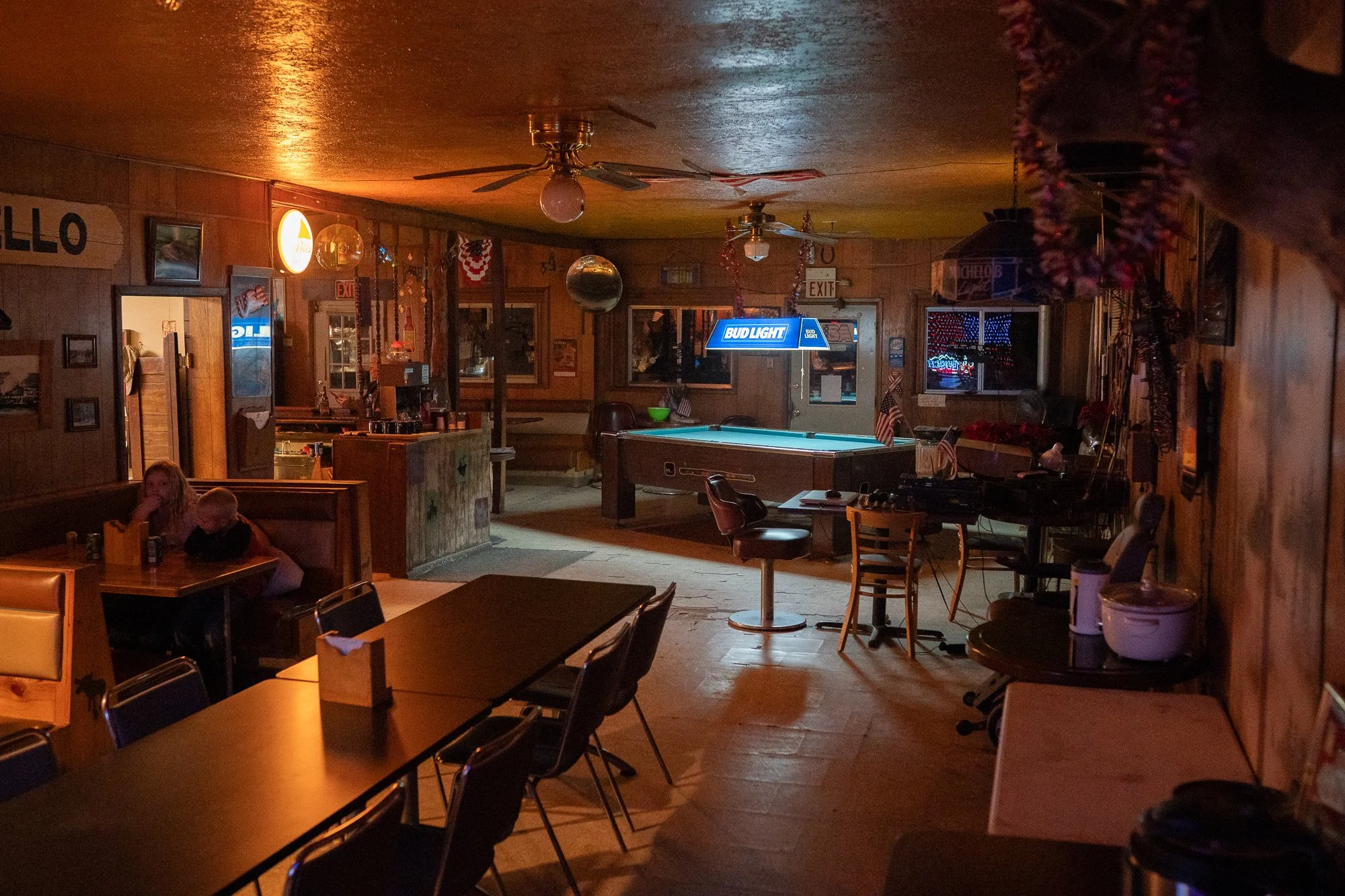 Dimly lit Nevada bar interior with wood-paneled walls, a pool table under a Bud Light lamp, vinyl booths, and one patron seated in the corner.