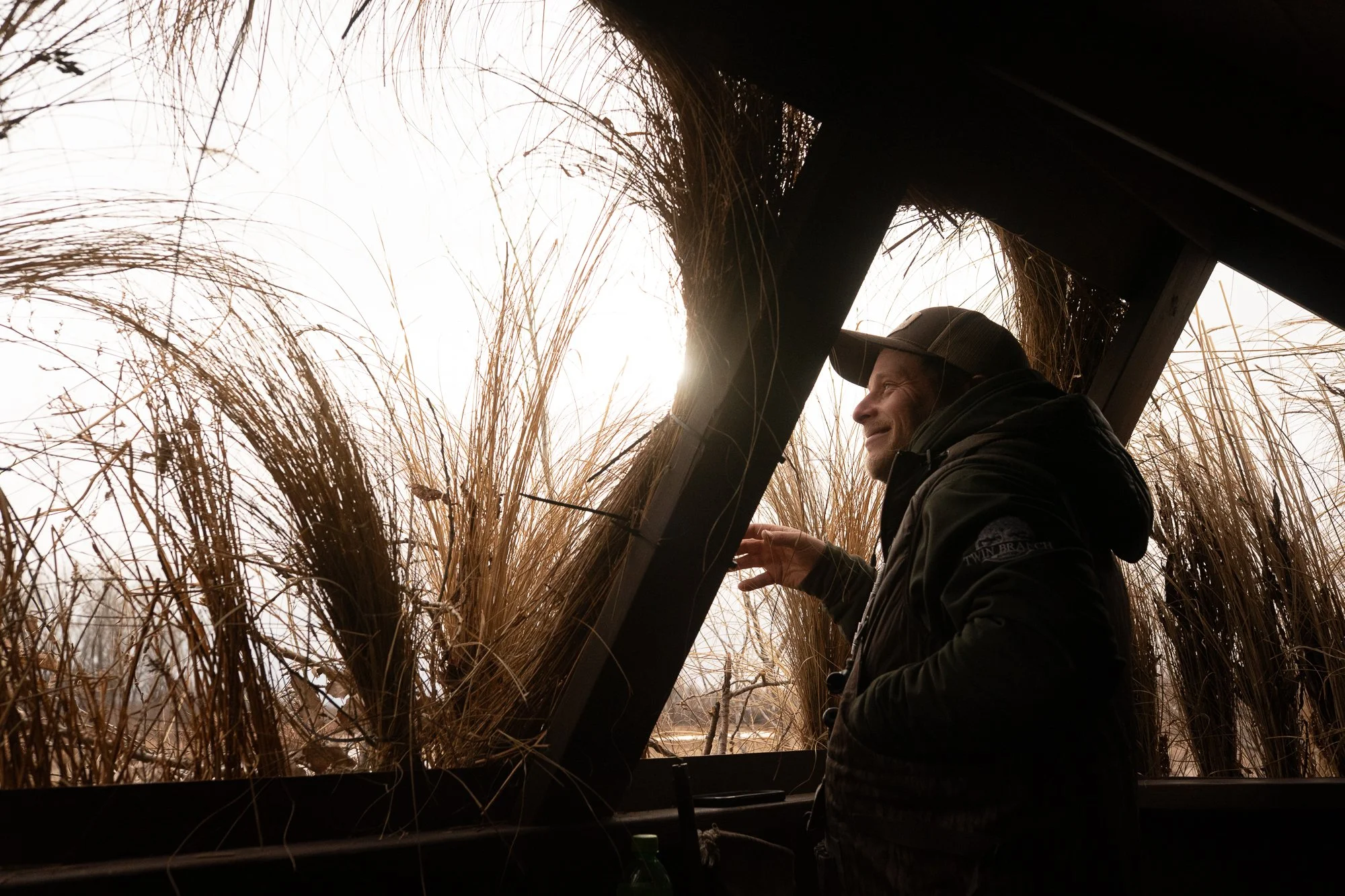 Duck hunters stand beside a grass-covered blind under headlamps before sunrise