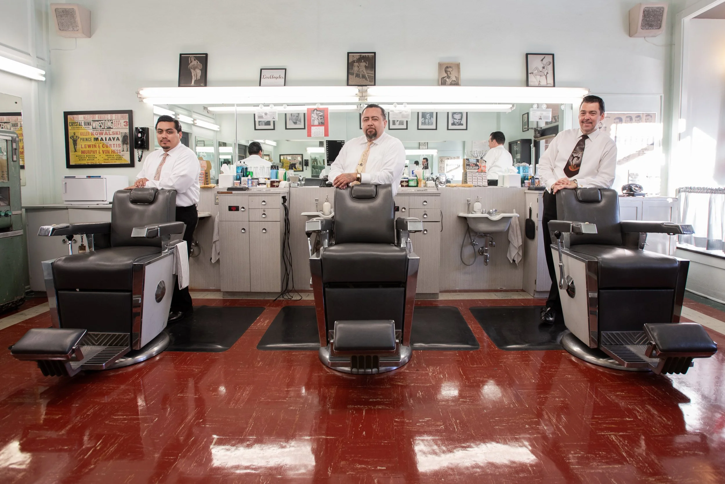 Three barbers in white shirts and ties standing behind three vintage barber chairs at Sweeney Todd's Barbershop, reflected in wall mirrors with vintage framed photographs and wrestling poster above