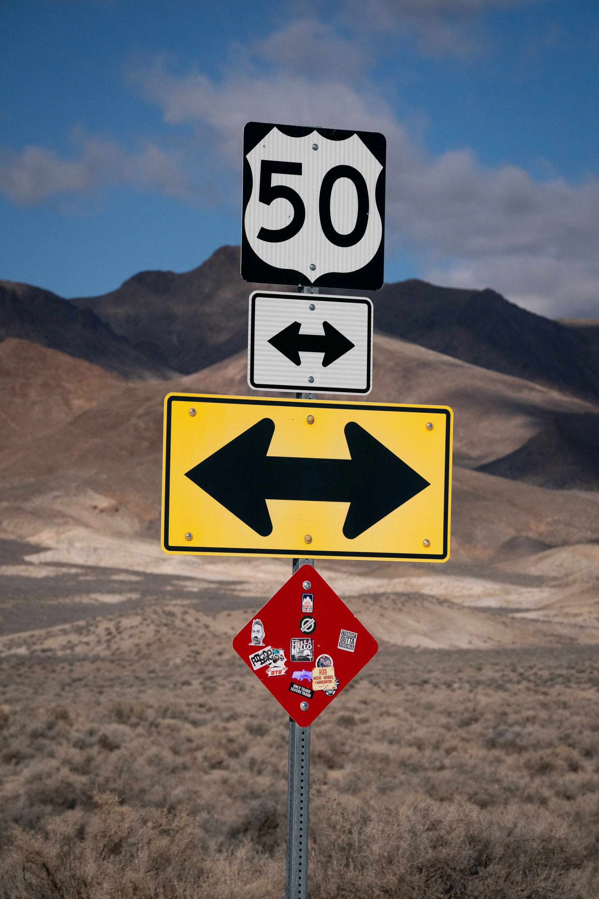 US Route 50 highway sign with directional arrows and a sticker-covered warning sign below it, set against a dry Nevada desert hillside under a partly cloudy sky.