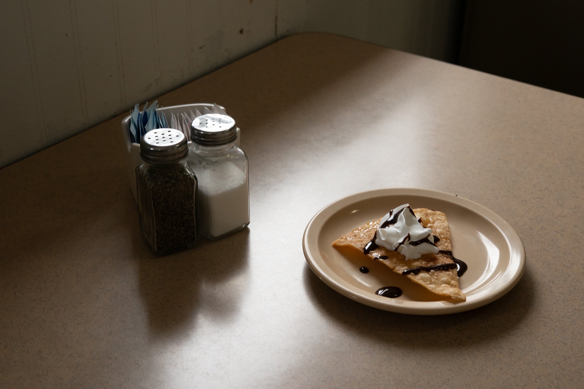 Slice of dessert with whipped cream and chocolate sauce on a plate beside salt and pepper shakers on a diner table.