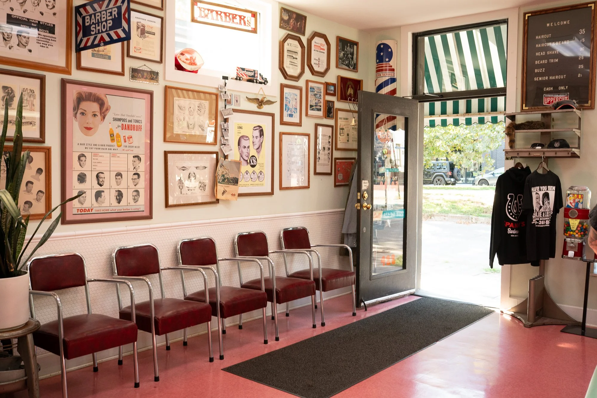 Waiting area inside Spanky’s Barbershop in Covington, Kentucky with red vinyl chairs, vintage barber posters, and green striped awning visible through the open door.