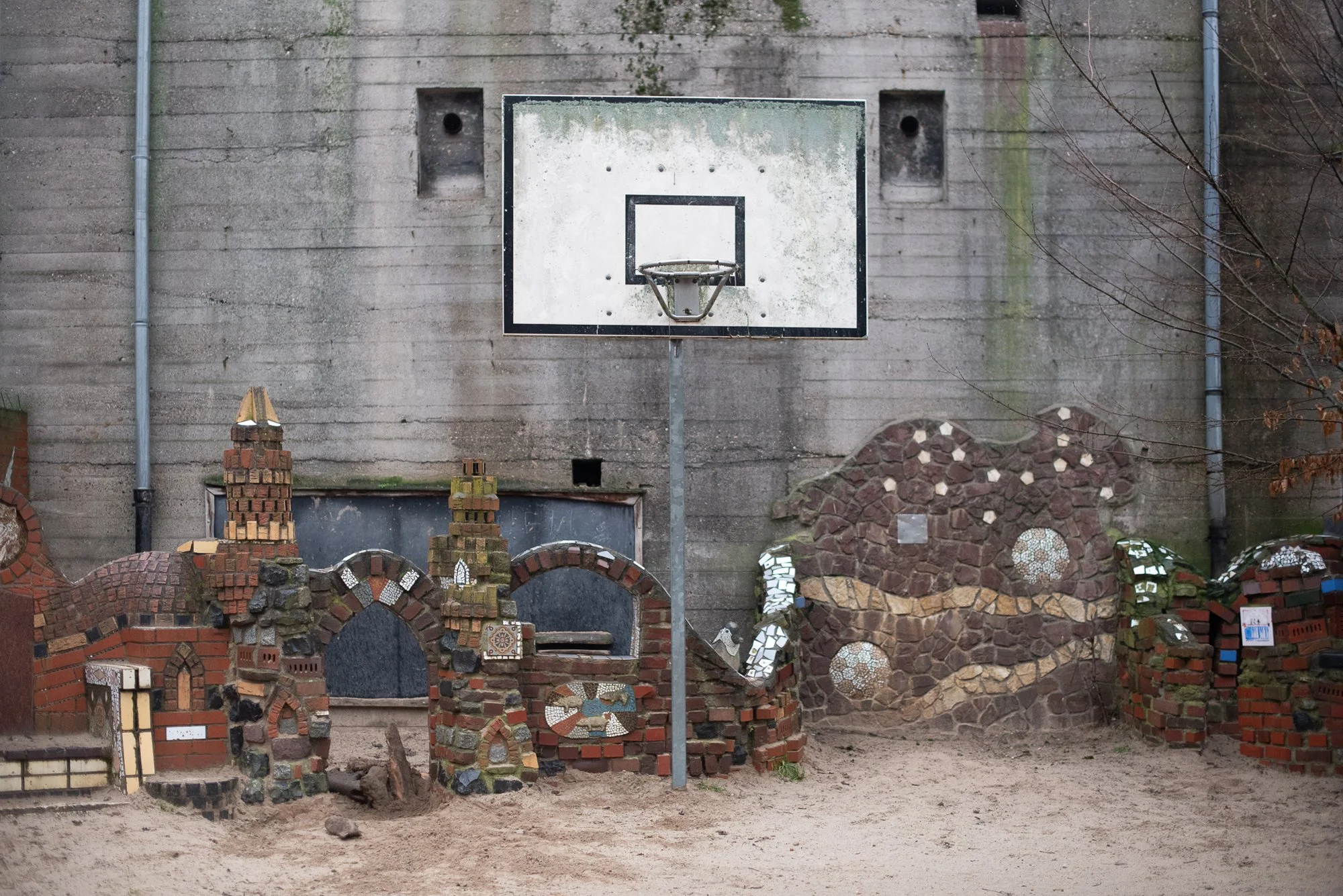 Outdoor basketball hoop on a metal pole set in front of a mosaic brick wall on a European courtyard court.