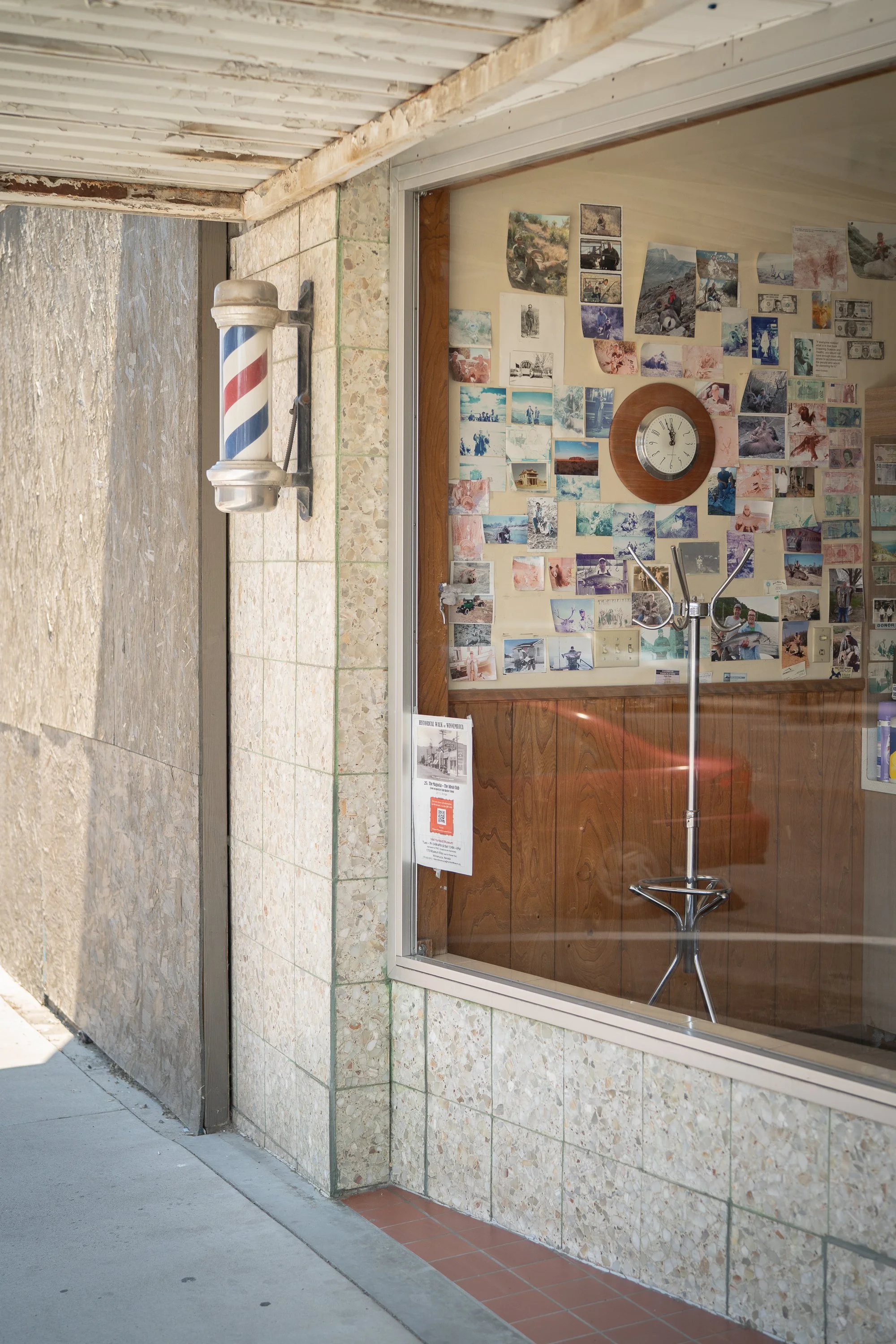 Small Nevada barbershop exterior with a mounted barber pole, photos covering the interior wall, and a wall clock visible through the front window.
