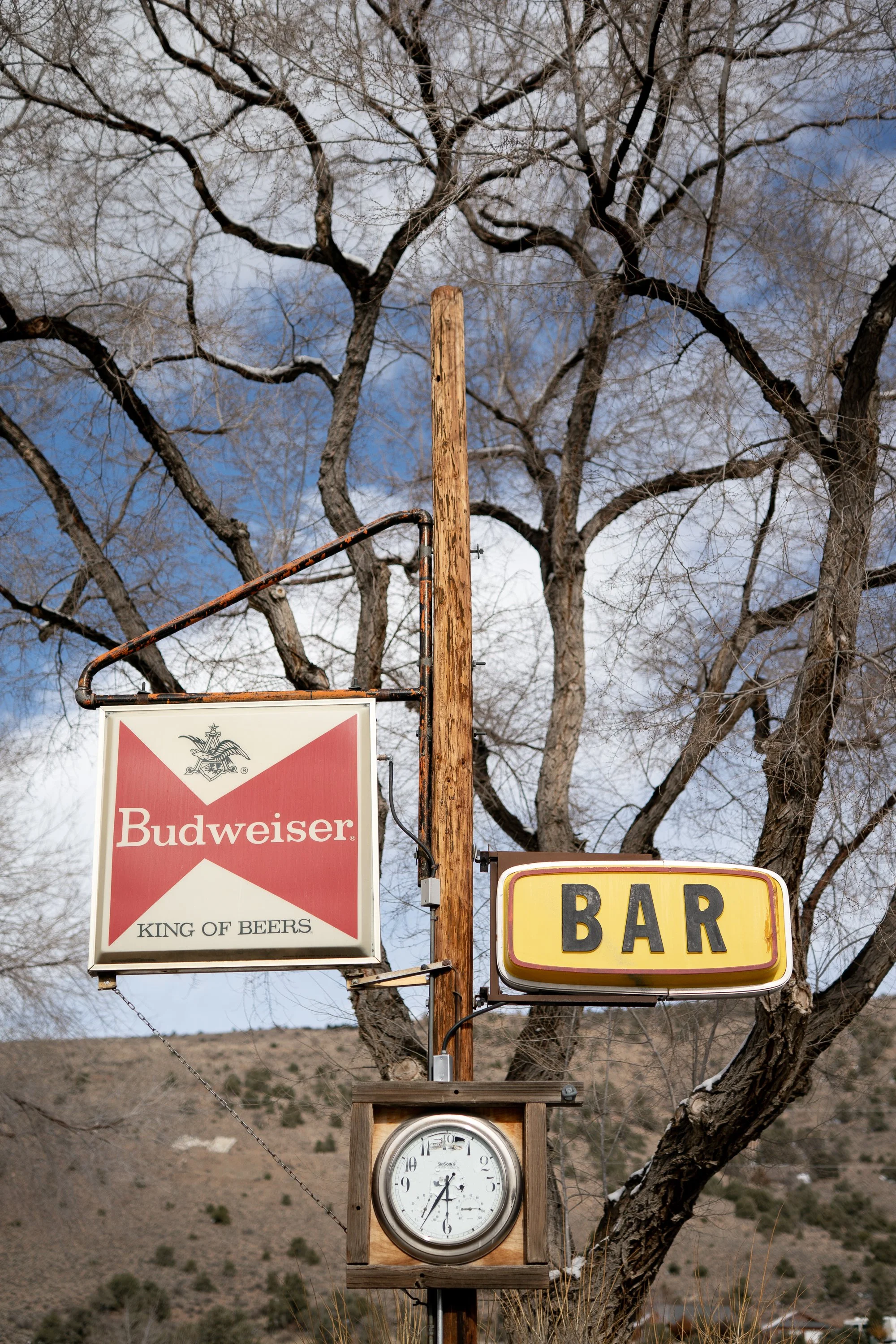 Vintage Budweiser King of Beers sign and a yellow Bar sign mounted on a wooden post, with a clock below and bare winter trees and Nevada hills in the background.