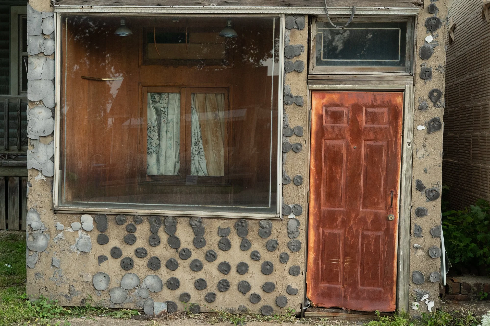 Building facade with rounded stones set into the exterior, a large front window with curtains, and a worn orange door.