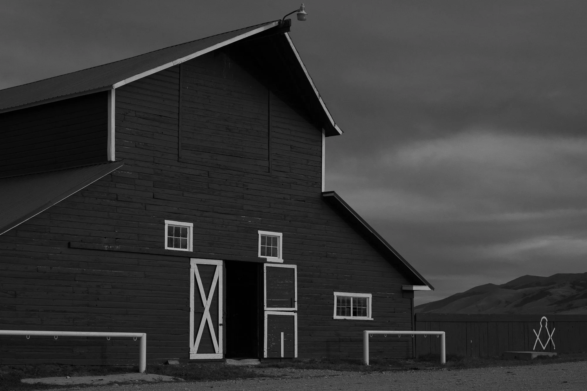 Black-and-white photograph of a large wooden ranch barn with white-trimmed doors and windows, a marked fence, and mountains beneath a cloudy sky.