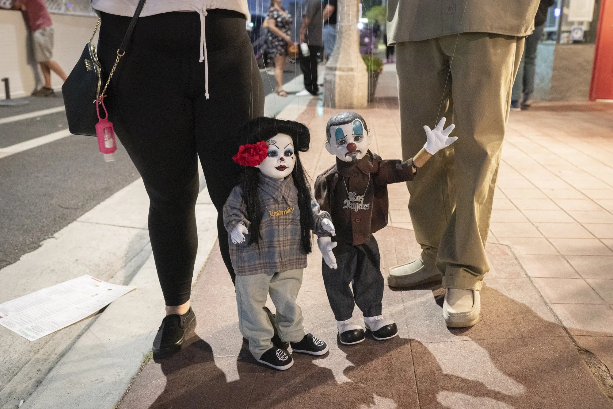 Two Chicano-style marionette puppets dressed in streetwear standing on the sidewalk between two people's legs during the Syndicate Barbershop anniversary event
