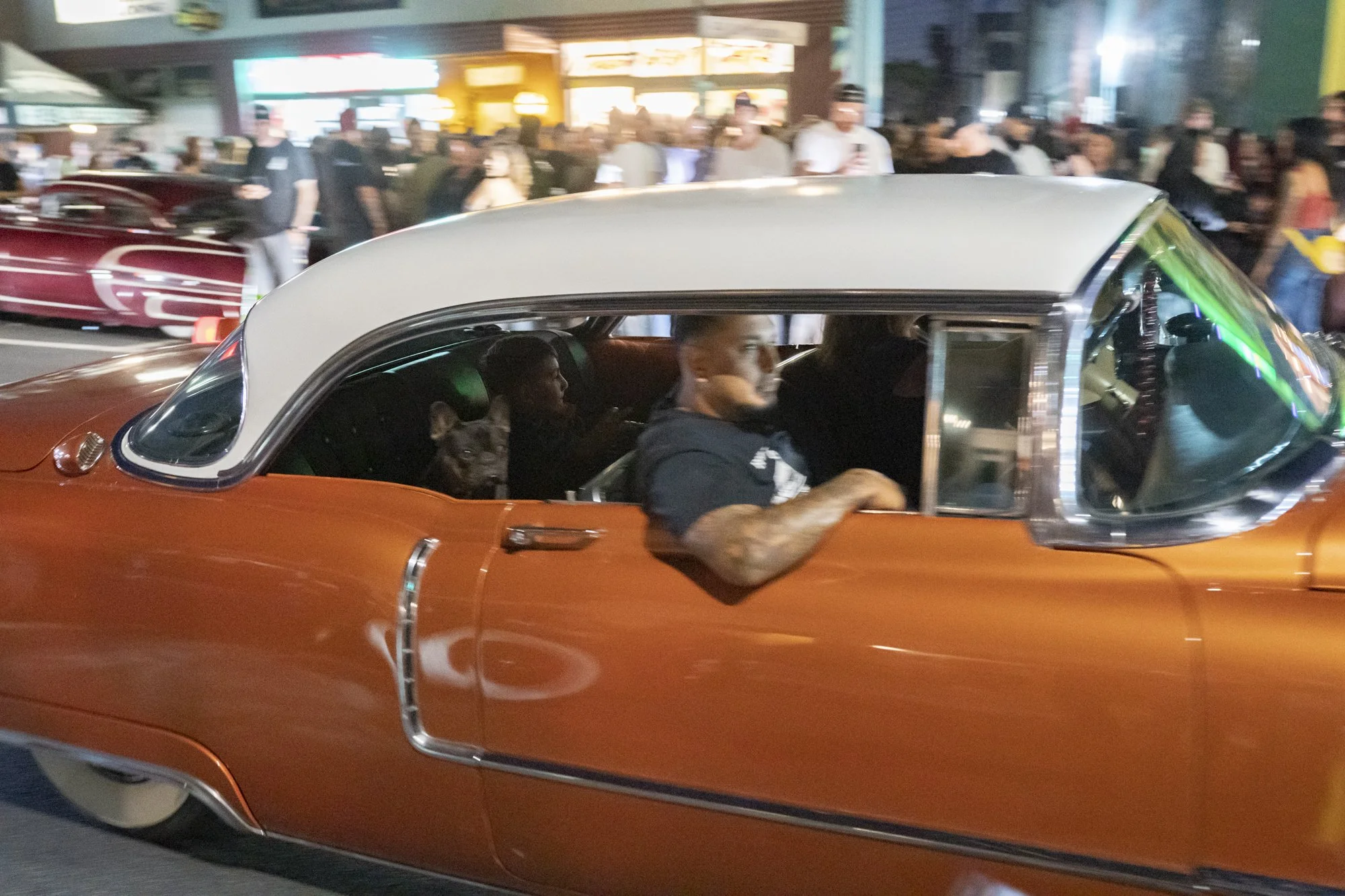 Copper-toned lowrider sedan with white roof moving through a crowd-lined Long Beach street at night during the Syndicate Barbershop 20th anniversary