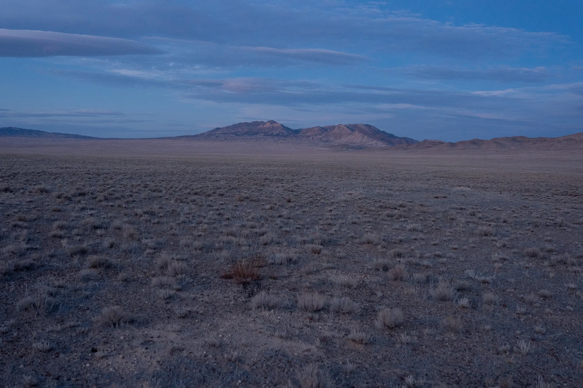 Wide flat Nevada desert plain covered in low sagebrush at early morning light, with a single isolated mountain range catching the first sunlight on the horizon.