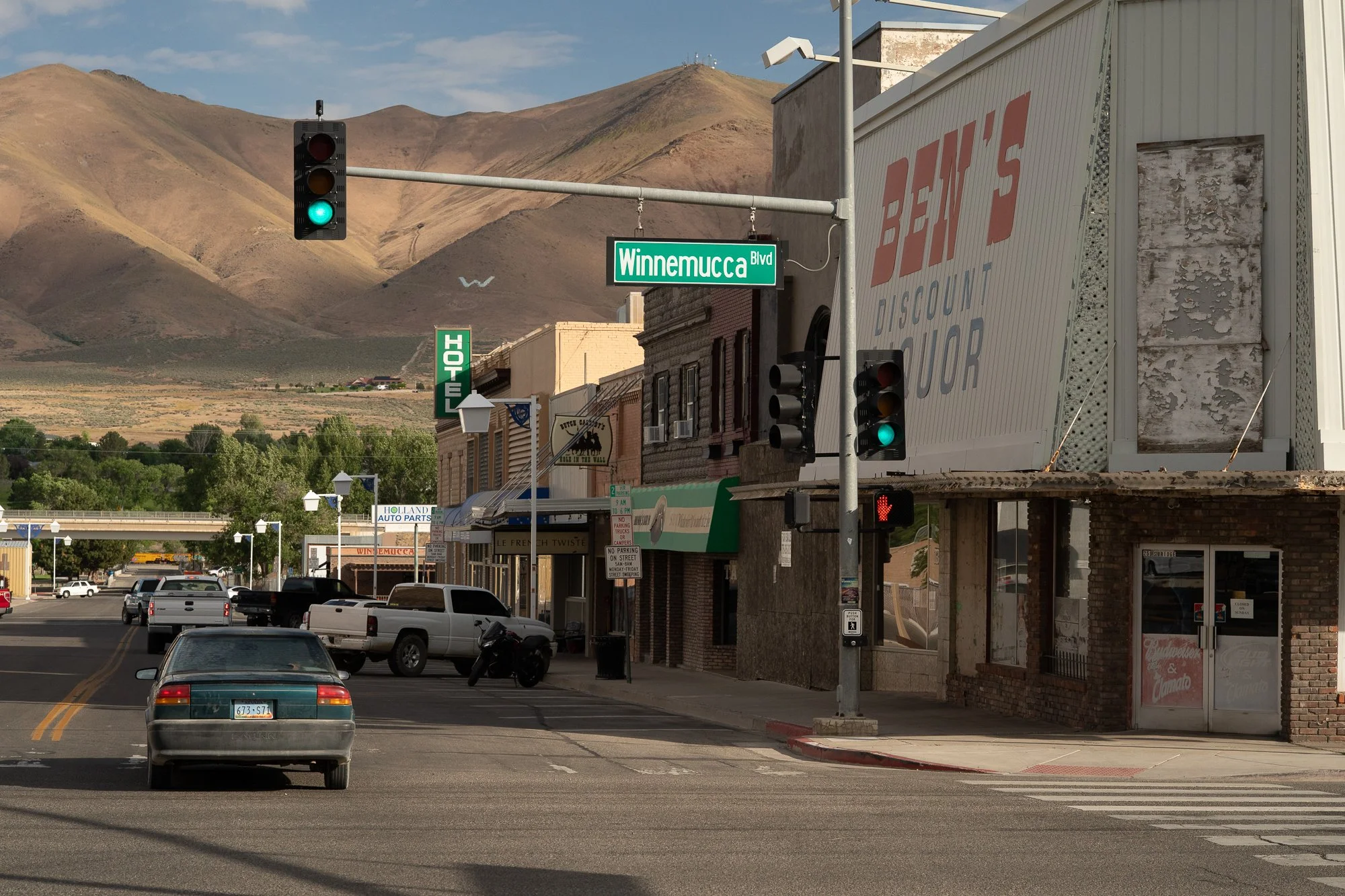 Winnemucca Boulevard in Winnemucca, Nevada with light traffic, a green street sign, Ben's Discount Liquor storefront, and a large desert mountain visible at the end of the street.