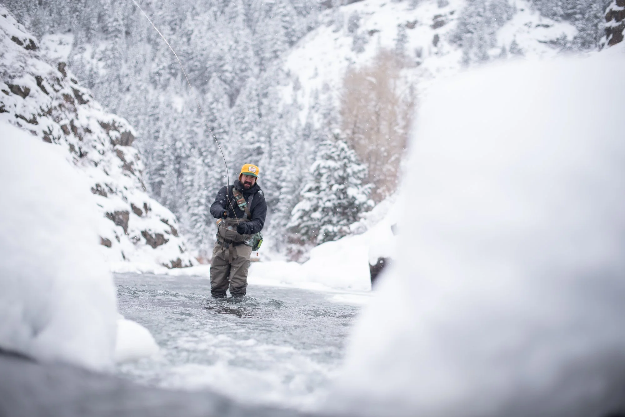 An angler casts a line while standing in Clear Creek during winter with snow falling around him.