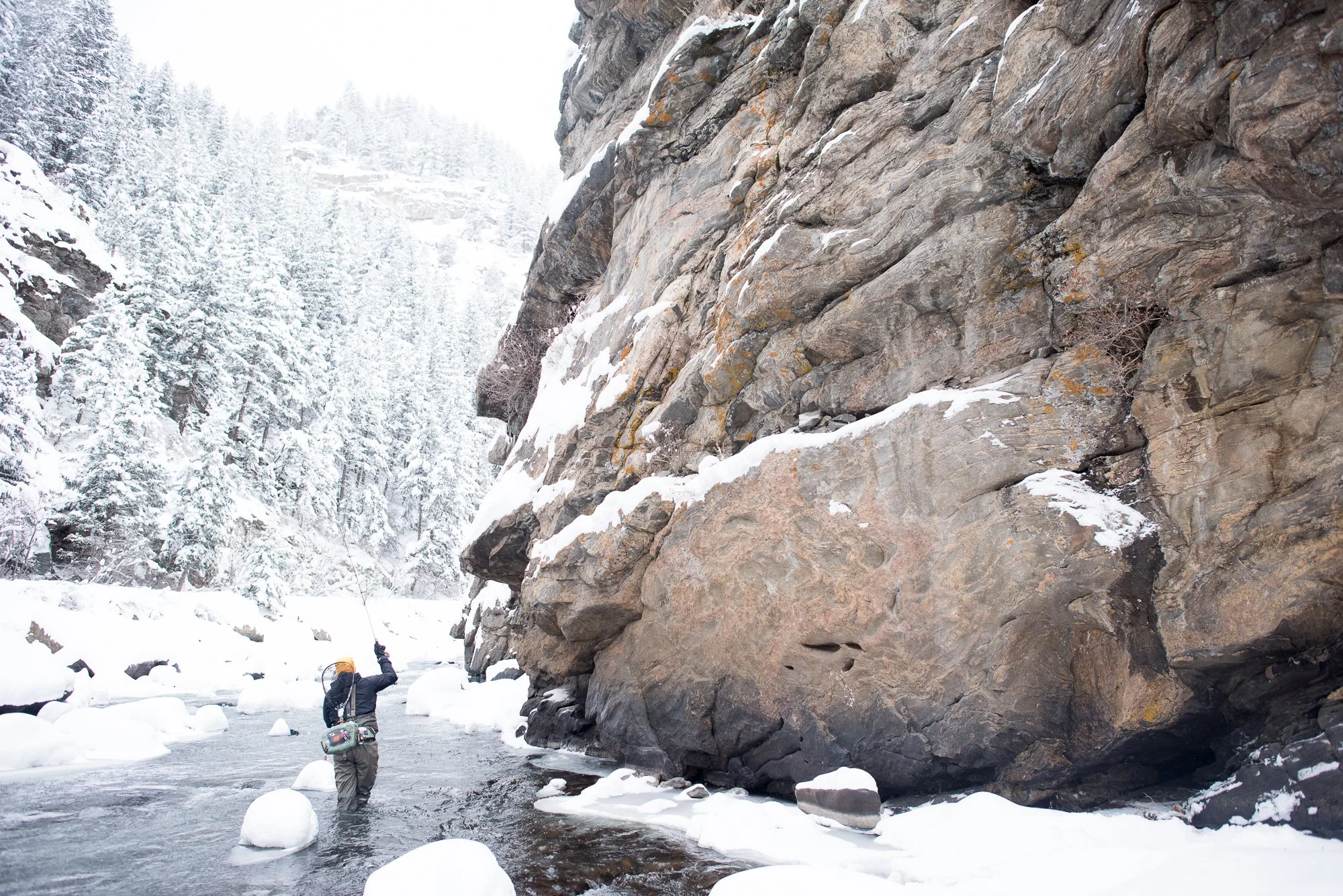 An angler fishes along a rocky wall on Clear Creek in Colorado during winter with snow-covered banks.
